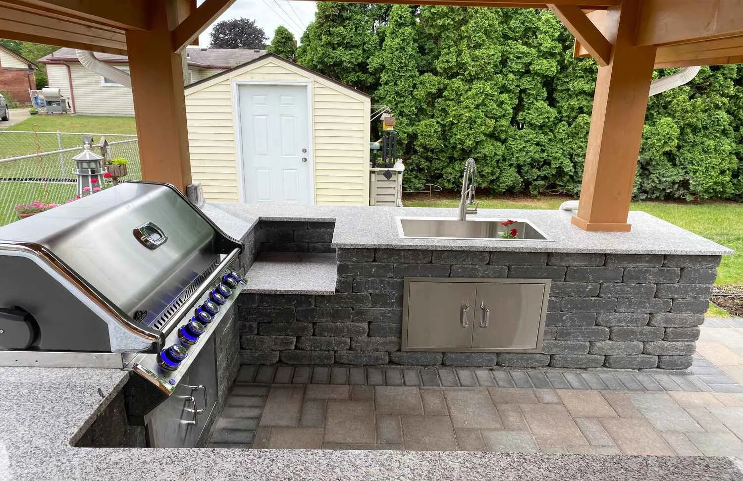 Outdoor kitchen with built-in grill, granite countertops, sink, and storage cabinet, surrounded by a backyard with a shed, trees, and a lawn.