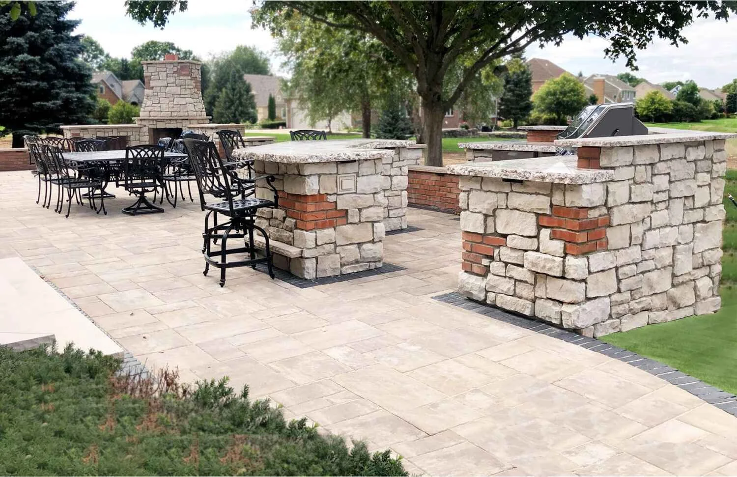 Outdoor patio area with stone and brick bar, grilling station, dining table, and chairs, surrounded by trees and neighborhood houses.