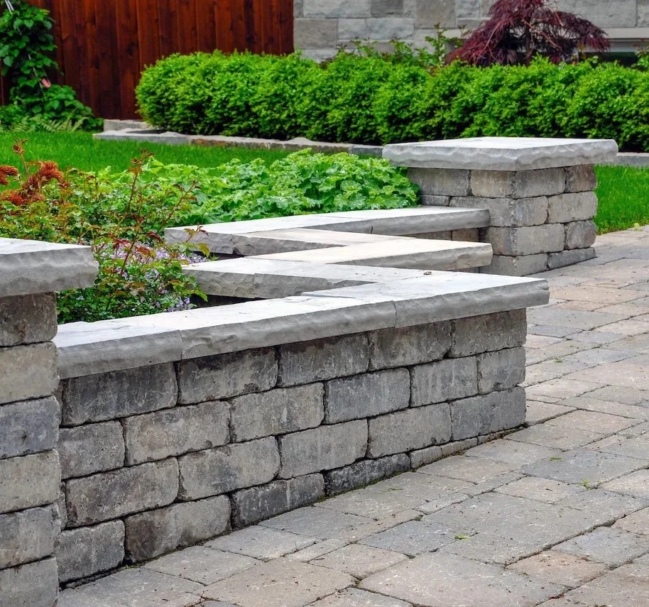 Stone garden bed with plants and bushes along a paved walkway
