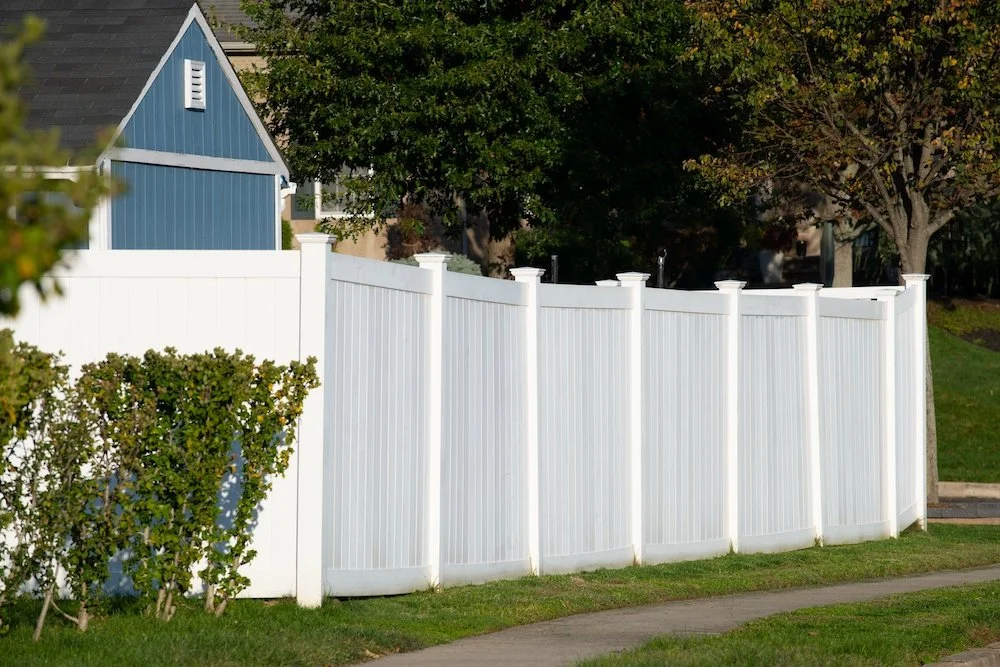 White privacy fence in front of trees and a small blue house with black roof.