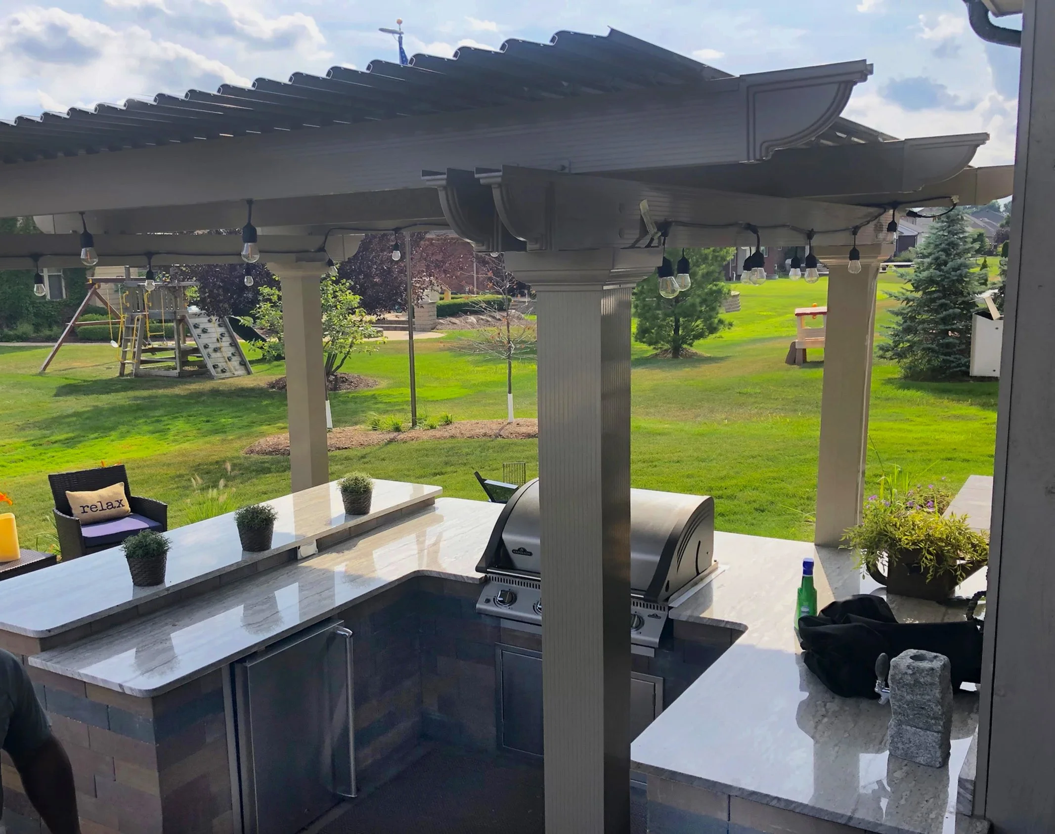 Outdoor bar area with a large marble countertop, a stainless steel grill, string lights, potted plants, and a grassy backyard with playground equipment and trees in the background.