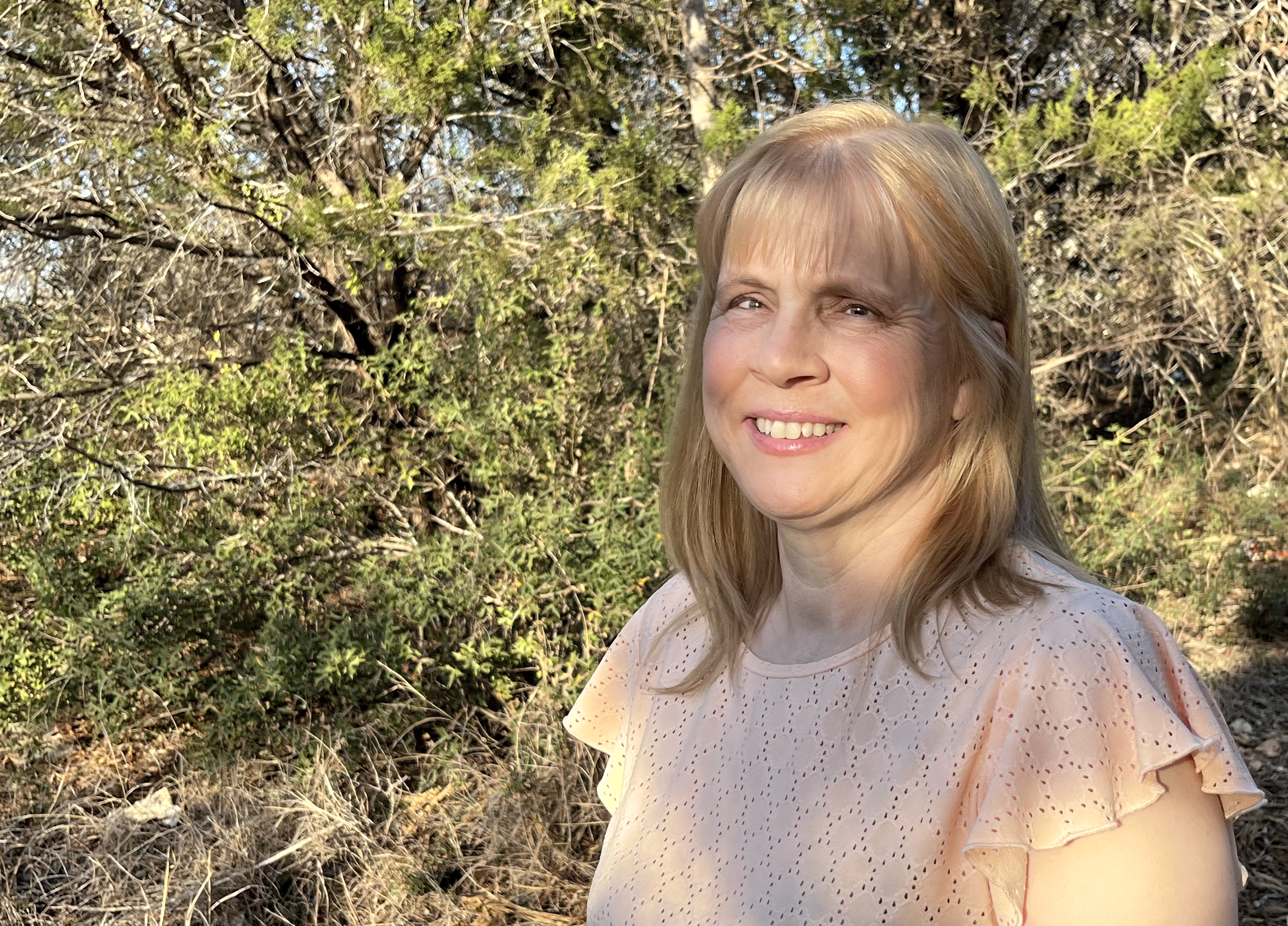 Image of Dawn Scott, blonde hair, light pink shirt. Outdoors, trees in background.