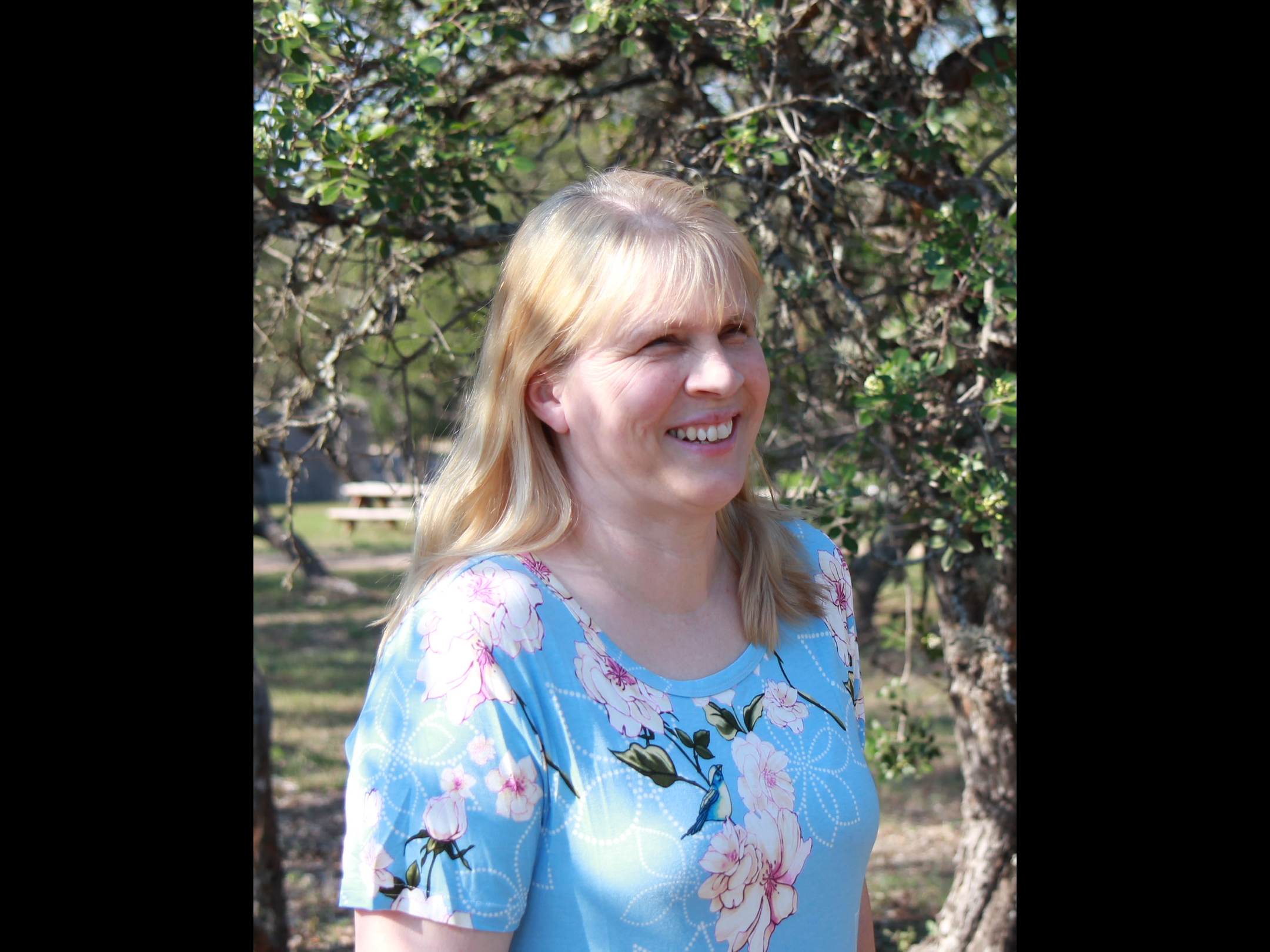 A woman with blonde hair smiling outdoors in a park or garden, with trees and green foliage in the background.