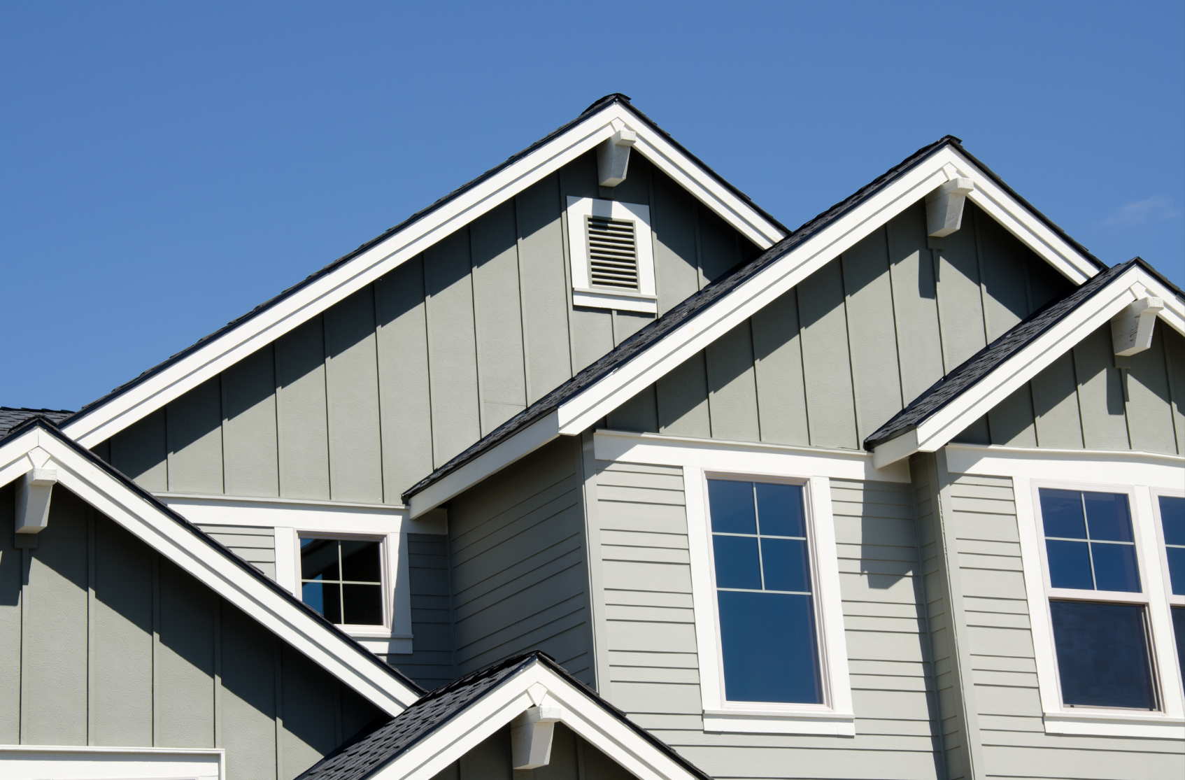 Multiple sections of a house with gray siding, gabled roofs, white trim, and windows against a clear blue sky.