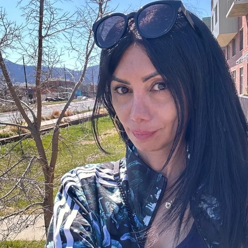 A woman with long black hair, wearing sunglasses on her head, hoop earrings, and a dark floral jacket, standing outdoors in a lavender field with trees in the background, during golden hour.