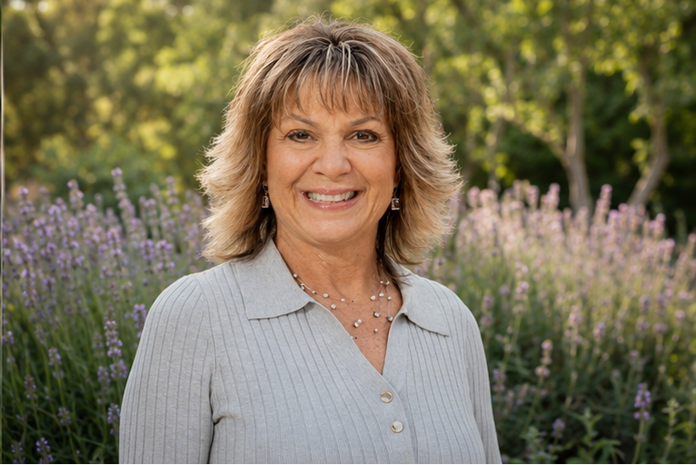 Smiling middle-aged woman with shoulder-length wavy hair, wearing a light-colored button-up shirt and jewelry, outdoors with sunlight and purple flowers in the background.