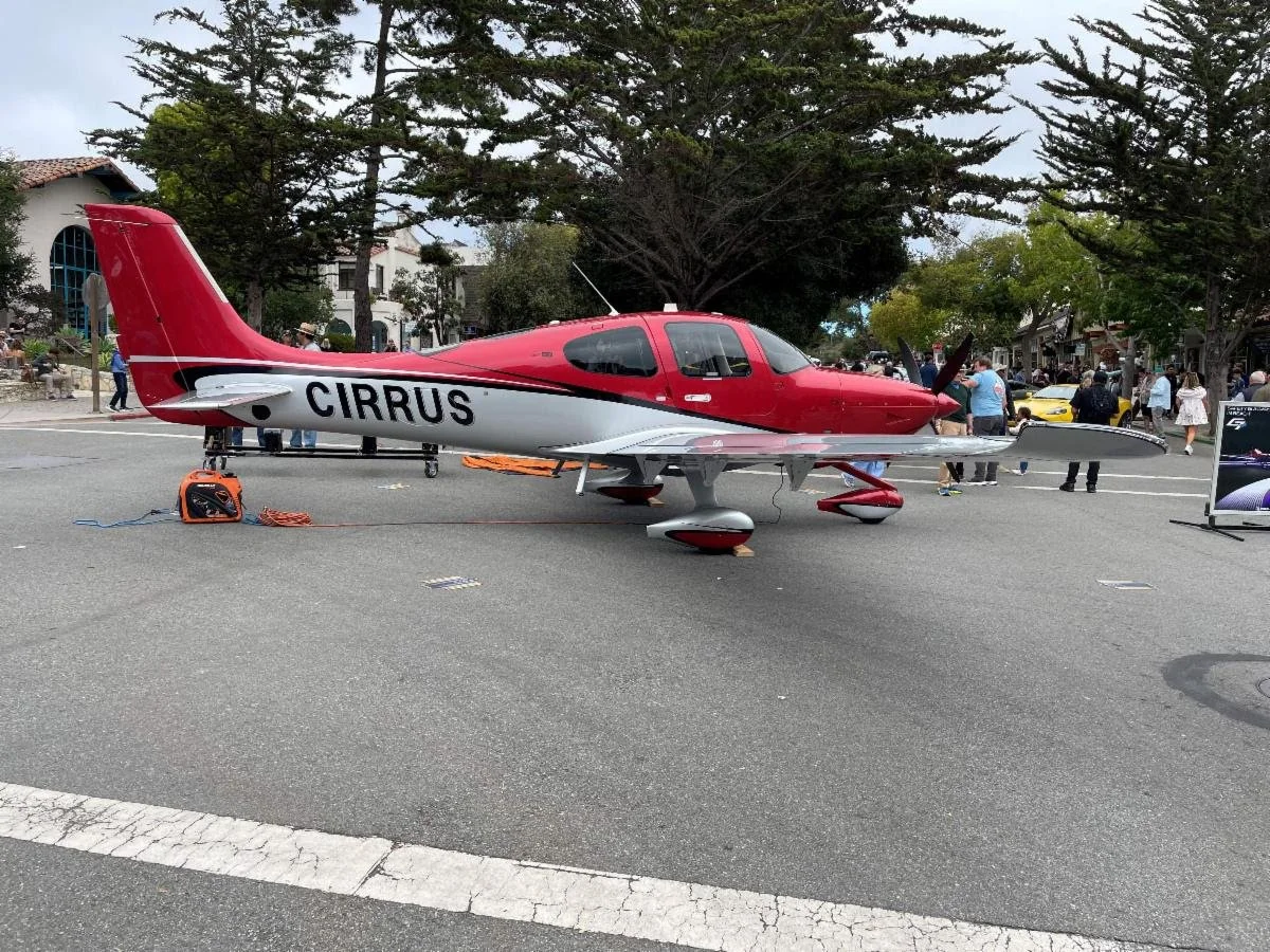 Red and white Cirrus aircraft displayed on a city street during an outdoor event, with people and cars in the background.
