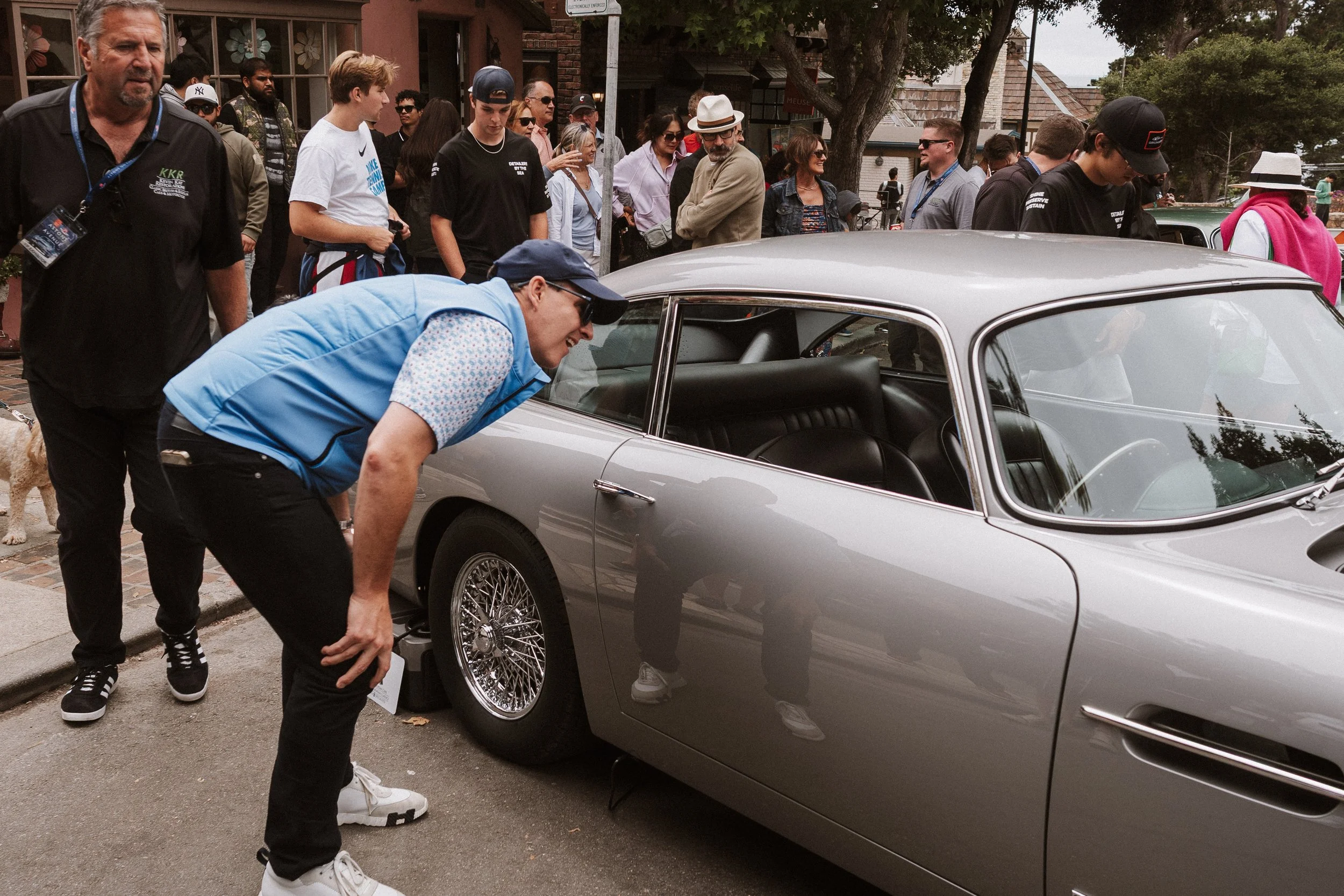 People gathered around a silver vintage car at an outdoor event, with a man in a blue vest inspecting the front left wheel.