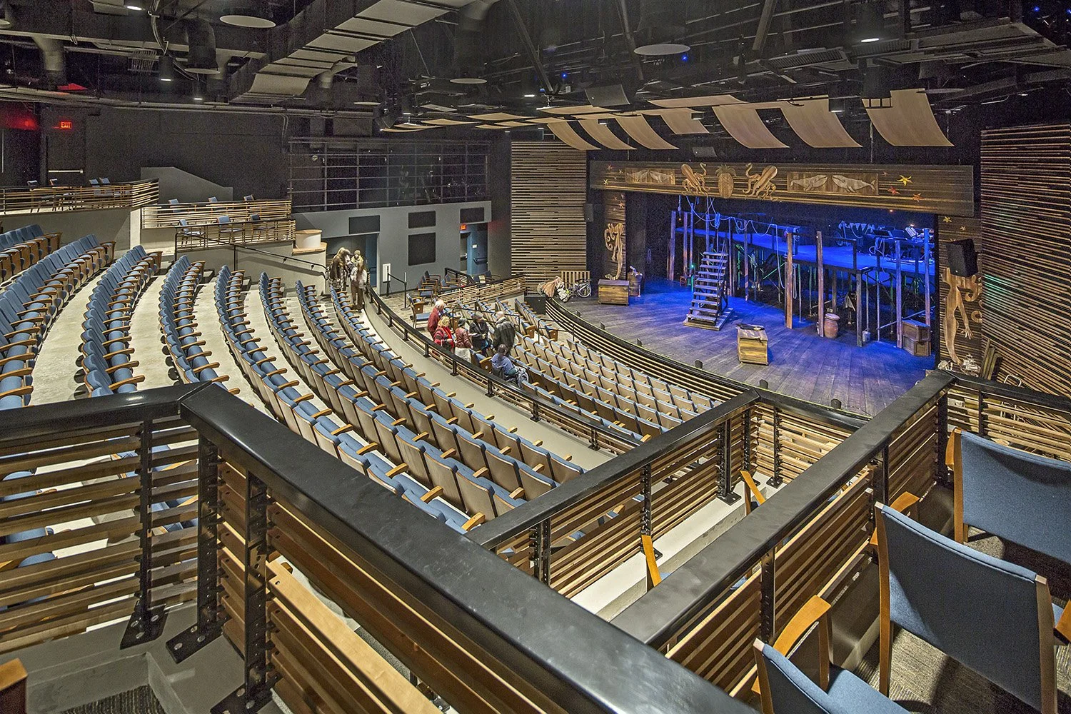 Empty theater with a small group of people near the stage, featuring wooden and blue seats and a stage with ropes and props.