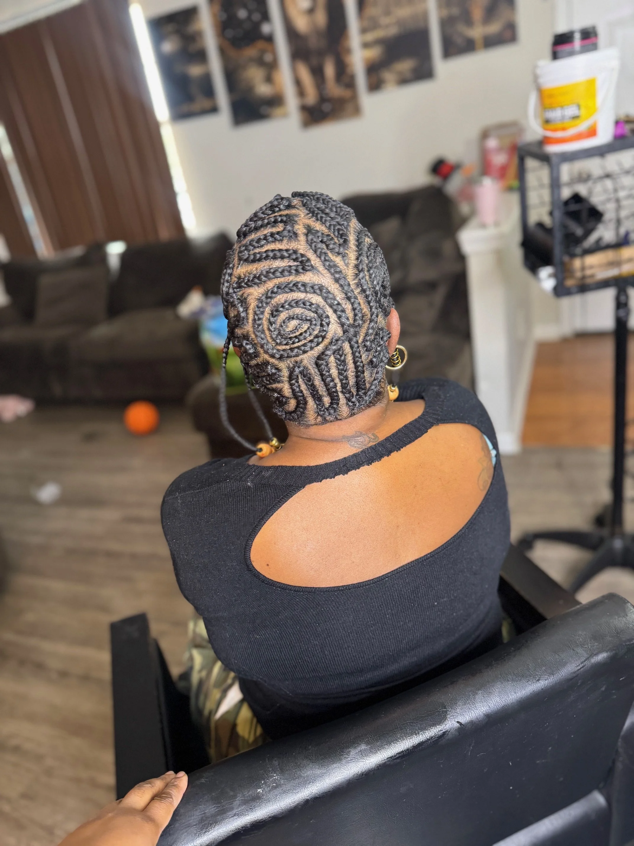 A woman with intricately styled hair featuring rosette and spiral patterns, seen from behind in a living room.