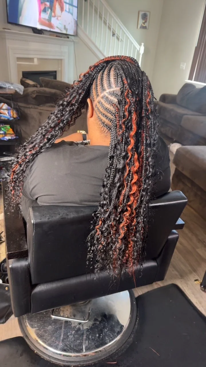 Person sitting in a salon chair with styled short twists with orange highlights, viewed from the back, in a living room with a TV and couch.