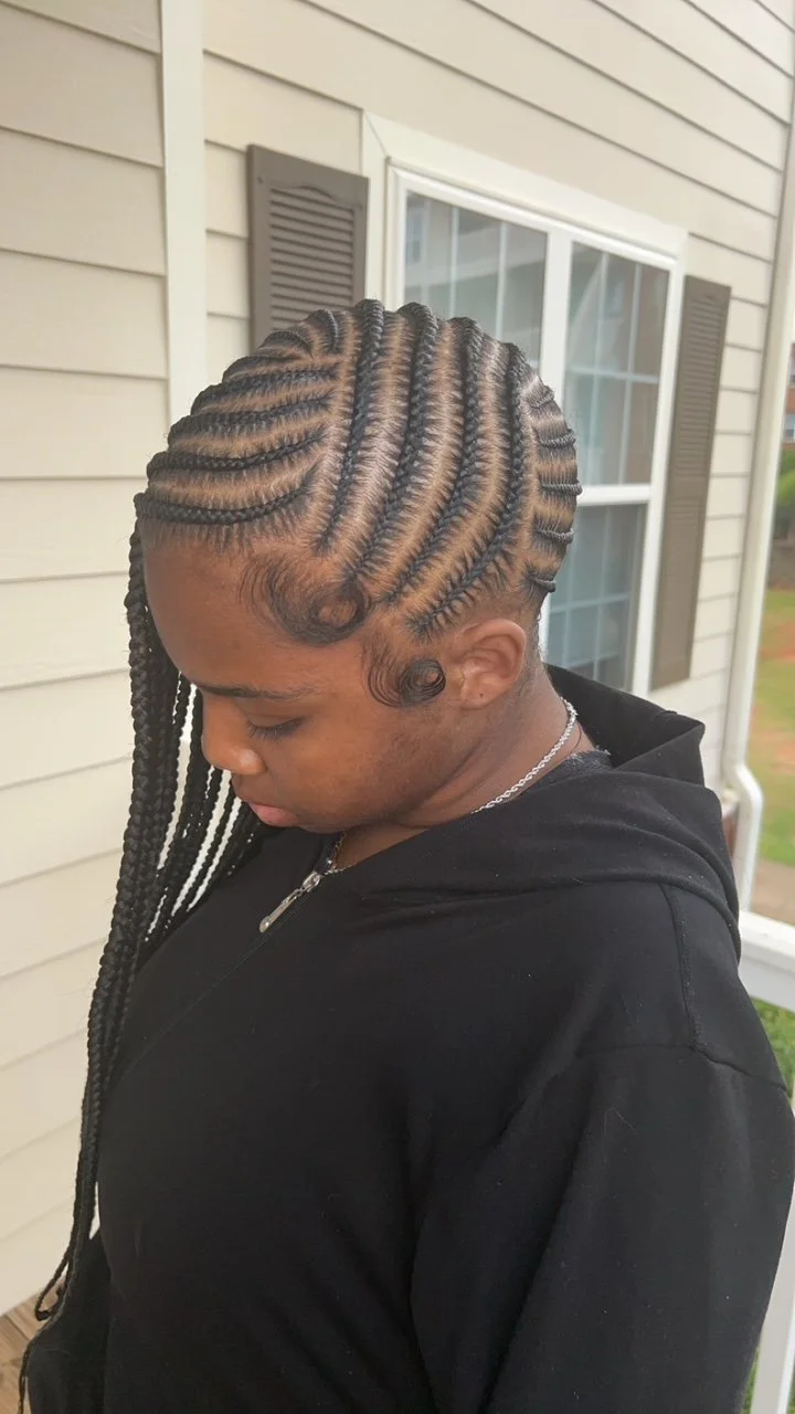 A woman with intricate, neatly braided hair styled in a pattern that sweeps toward her face, standing outside near a house with beige siding and a window with brown shutters.
