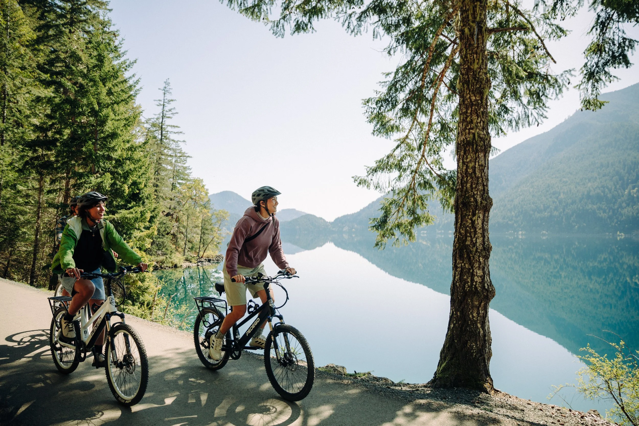 Two people riding bikes along a forested trail next to a lake with mountains in the background.