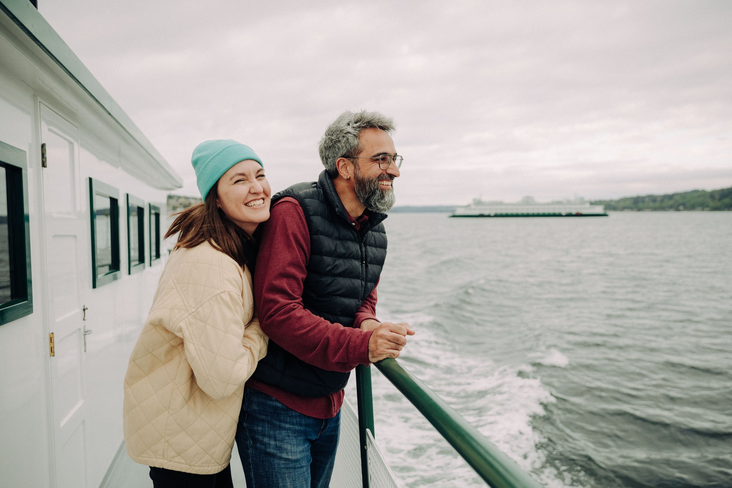 A smiling woman in a blue beanie and beige jacket hugs a smiling man with glasses and gray hair, wearing a black puffer vest and red shirt, as they stand on the deck of a boat looking out at the water with a distant ship and green shoreline.