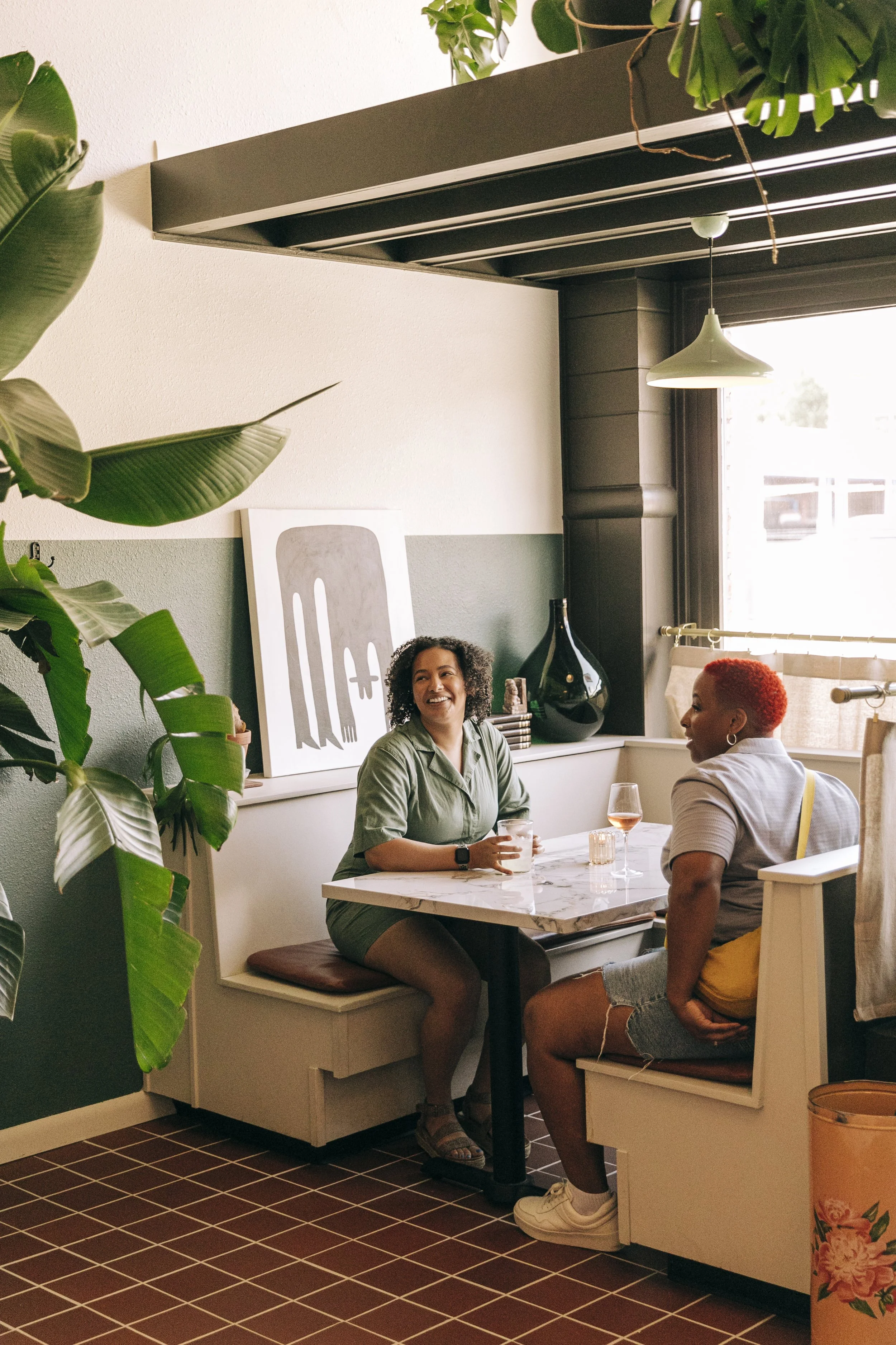Two women sitting in a cozy restaurant booth, smiling and having a conversation. One has curly hair and is holding a drink, the other has short red hair and is wearing a light-colored shirt. The restaurant has modern decor with artwork, plants, and a window letting in natural light.