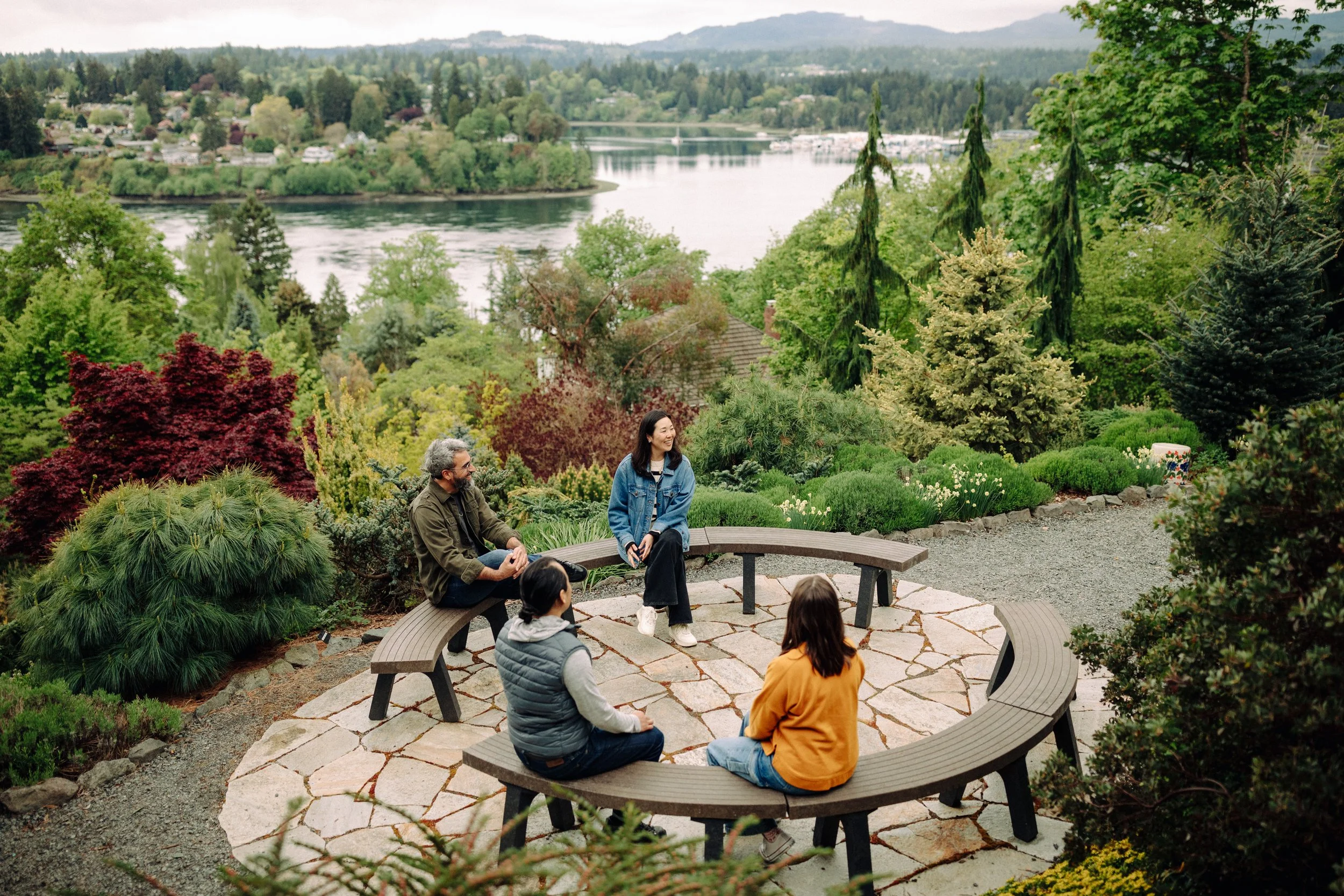 A group of five people sitting on circular benches in a well-maintained garden overlooking a lake and lush green trees.