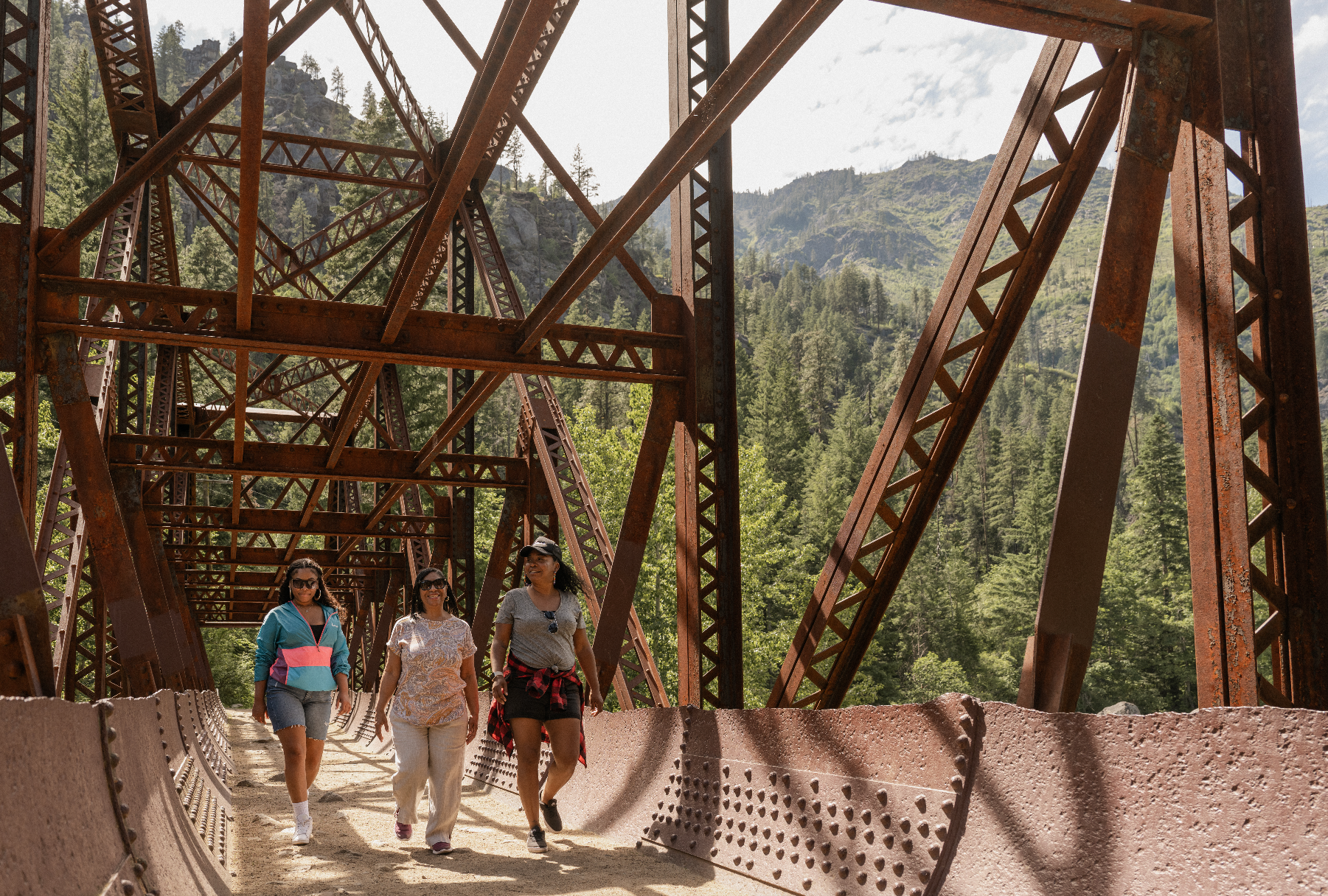 Three women walking on a rusty iron bridge surrounded by a forested mountainous landscape.