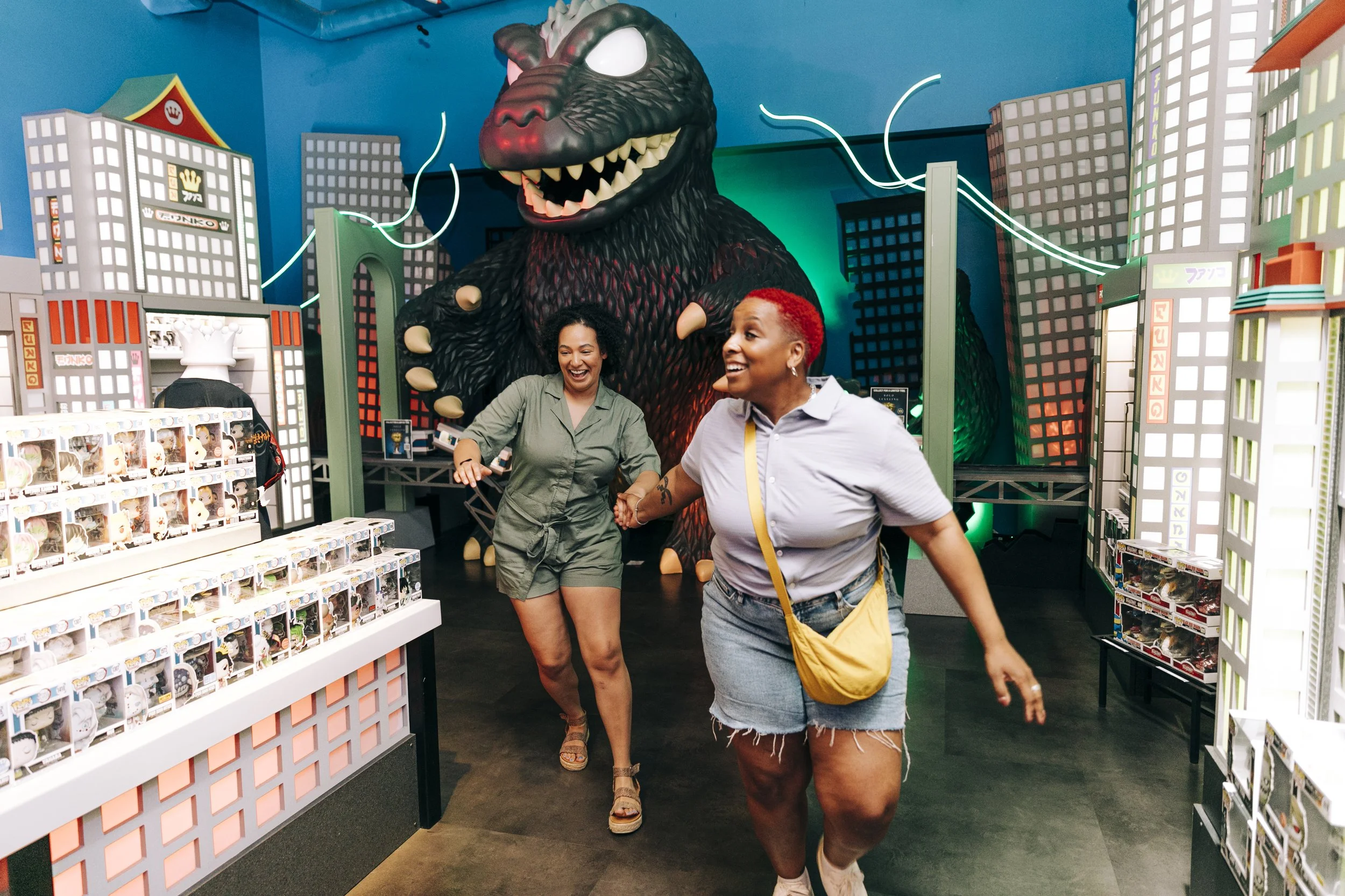Two women are holding hands and enjoying their time in a toy store with a large Godzilla figure and cityscape decorations in the background.