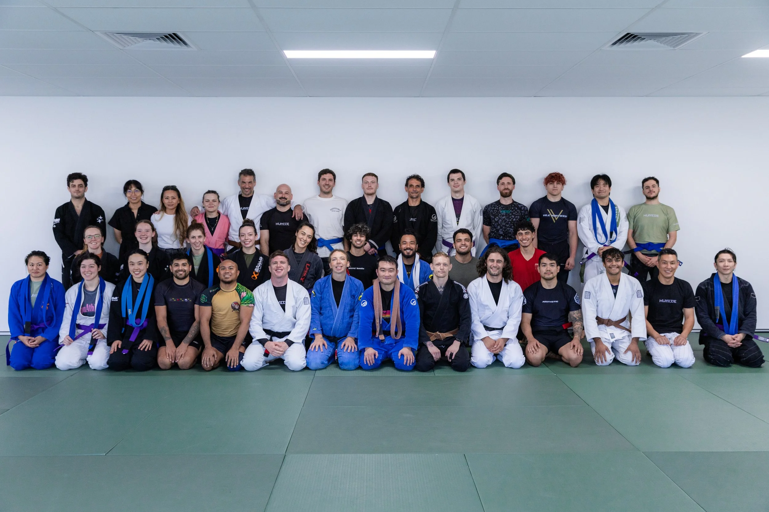 Group photo of martial artists and instructors in uniform, sitting and standing on a matted training floor, with a plain white wall in the background.