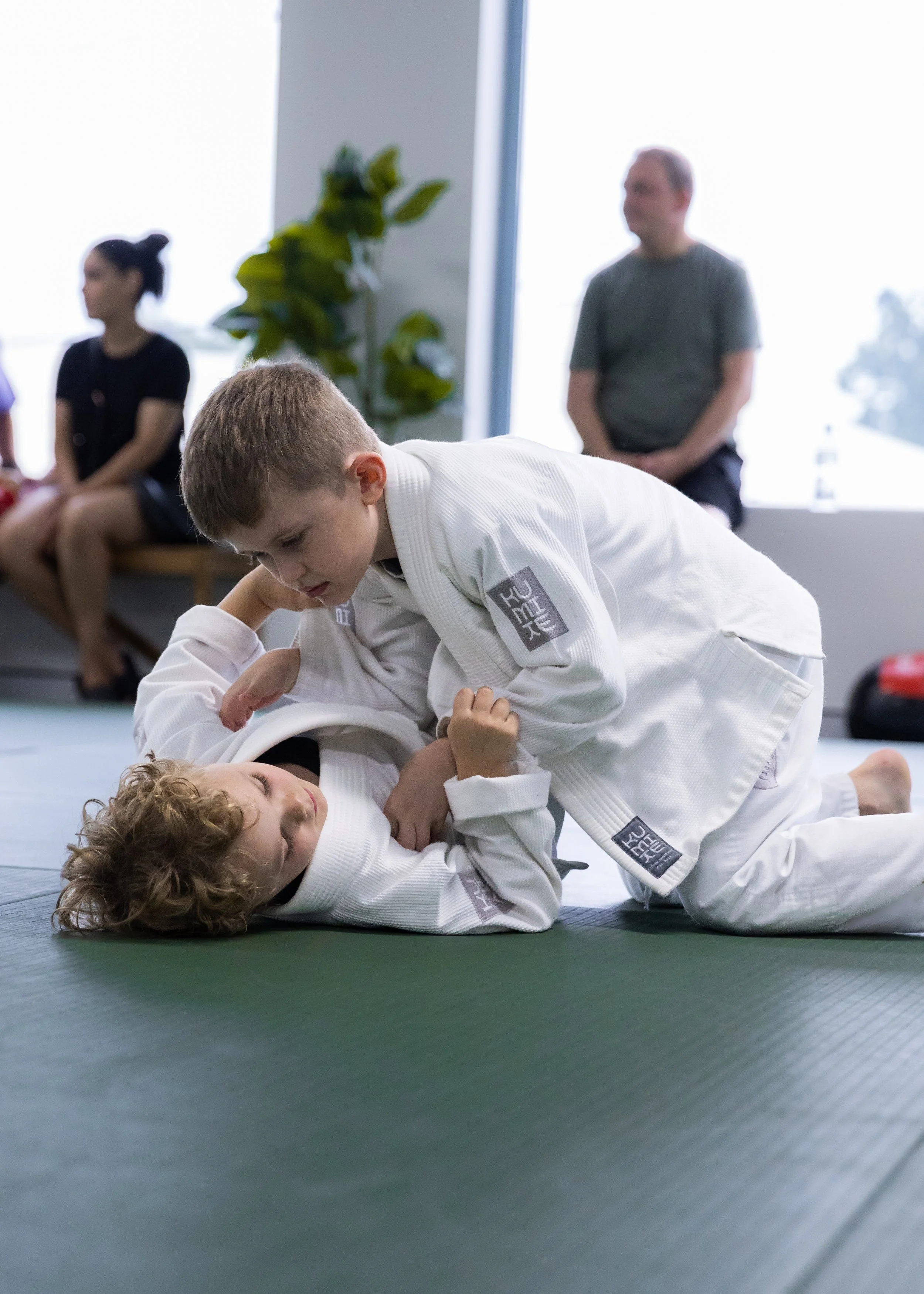 Two children practicing Brazilian Jiu-Jitsu on a mat, with one child on top and the other on the ground gripping the top child's arm, in a martial arts class setting with an adult and other children watching in the background.