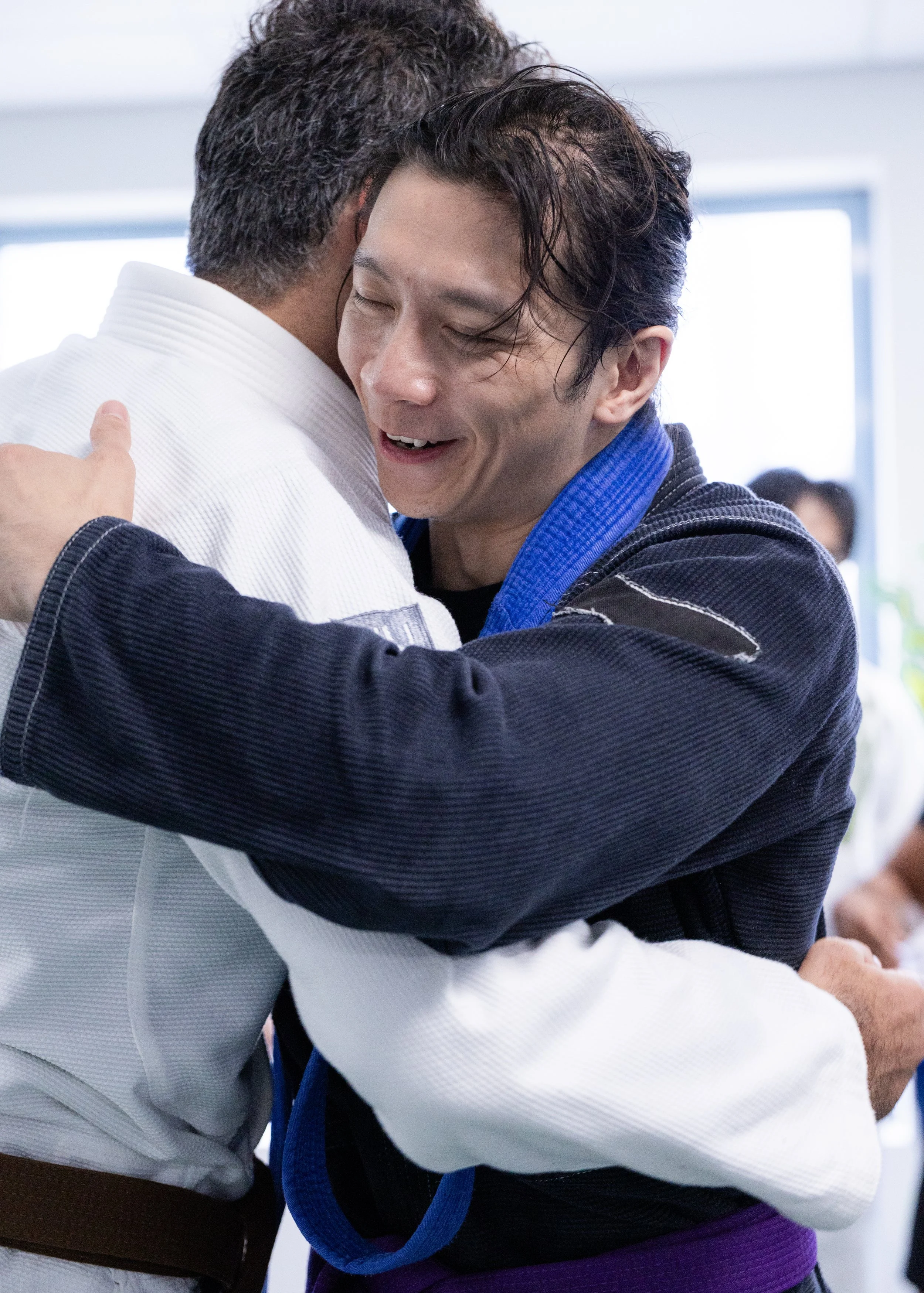 Two men hugging, one in a judo gi and the other in a black jacket with a blue belt, smiling.