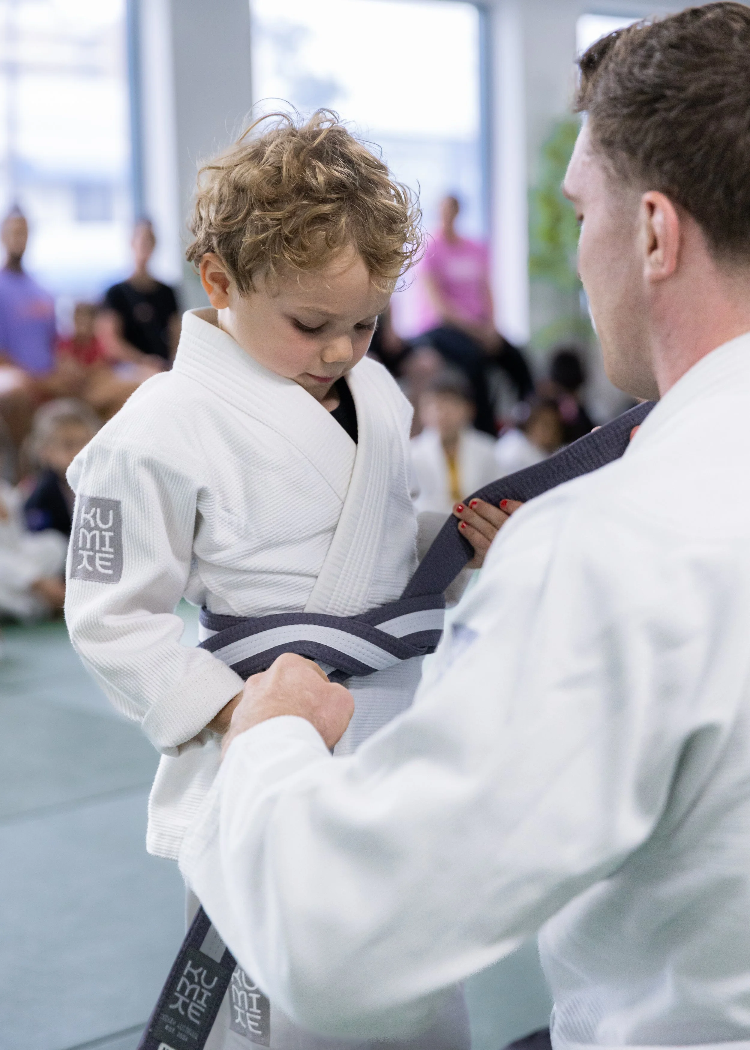 A young boy with curly hair wearing a white Judo gi getting a black belt tied by an instructor during a martial arts class or belt promotion ceremony, with other students watching in the background.