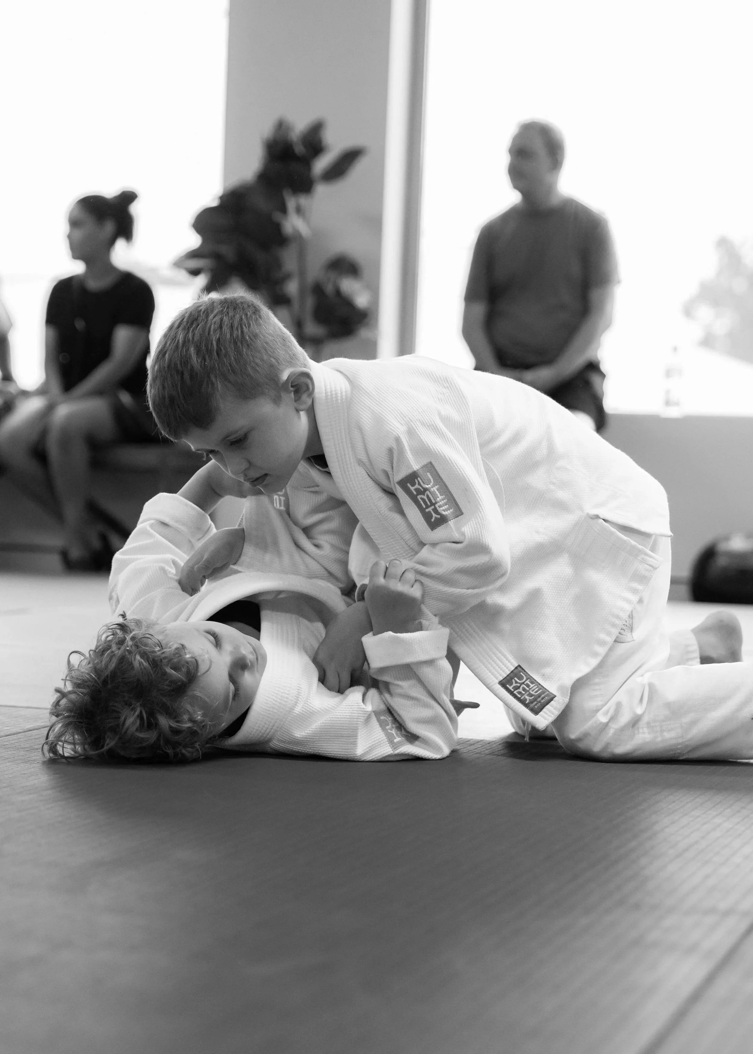 Two children practicing Brazilian Jiu-Jitsu on a mat, with one child in a white gi on the ground and the other child in a white gi on top, gripping each other's uniforms.