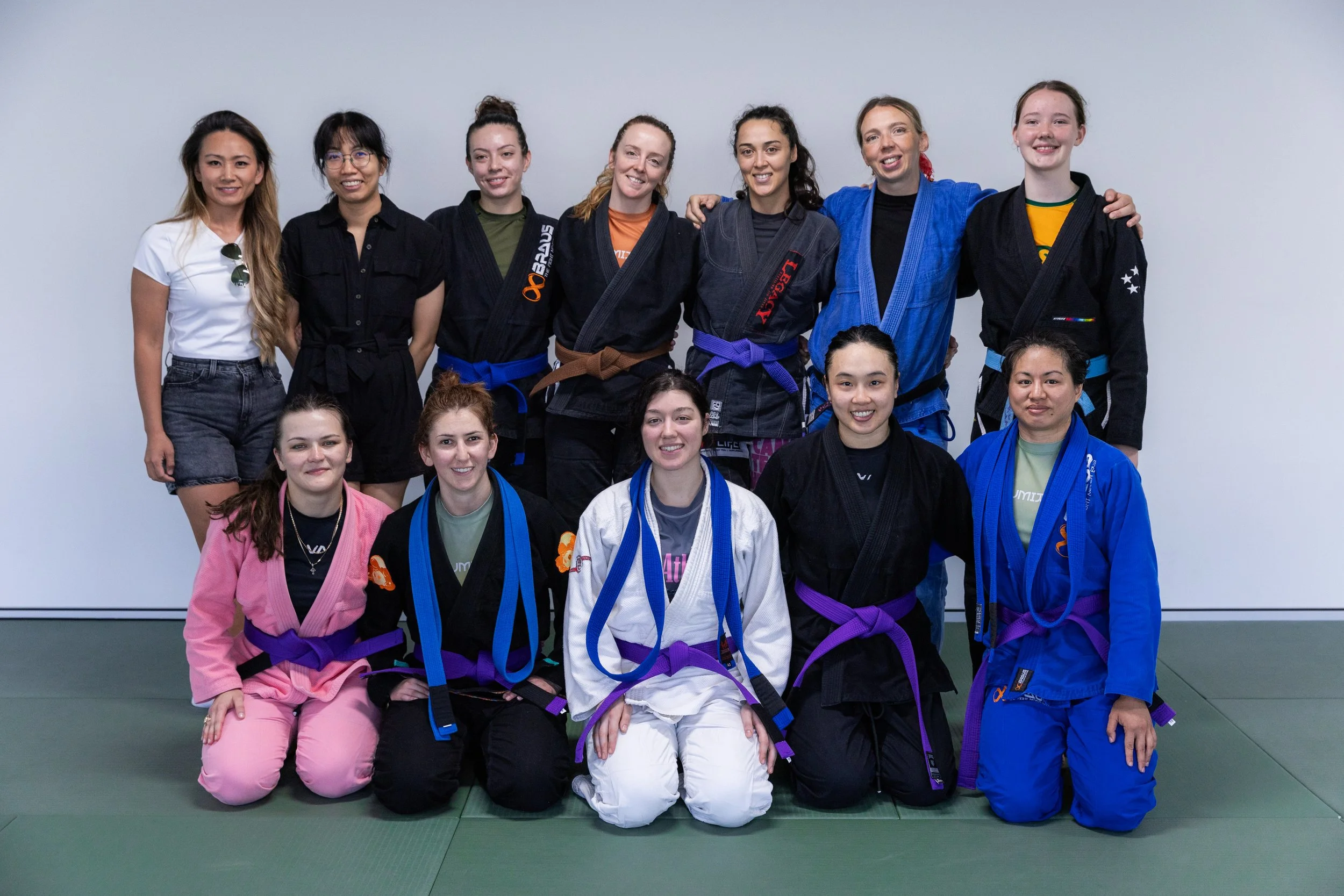 Group of 11 women in Brazilian Jiu-Jitsu gi uniforms and casual clothing, posing for a photo on the mat in a martial arts gym.