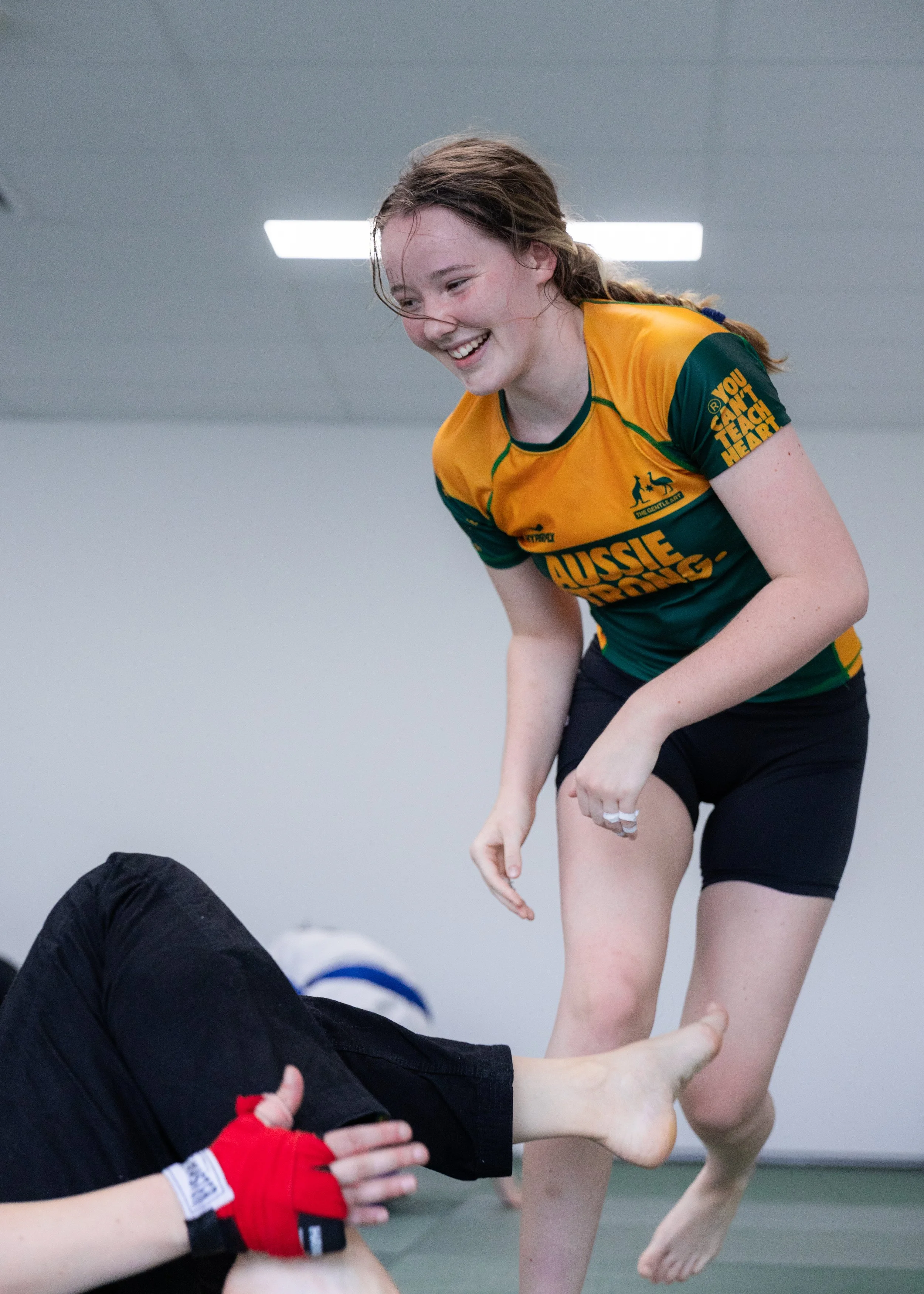 Young woman in yellow and green sports uniform playing combat sport in gym, smiling and leaning forward, with opponent lying on ground.