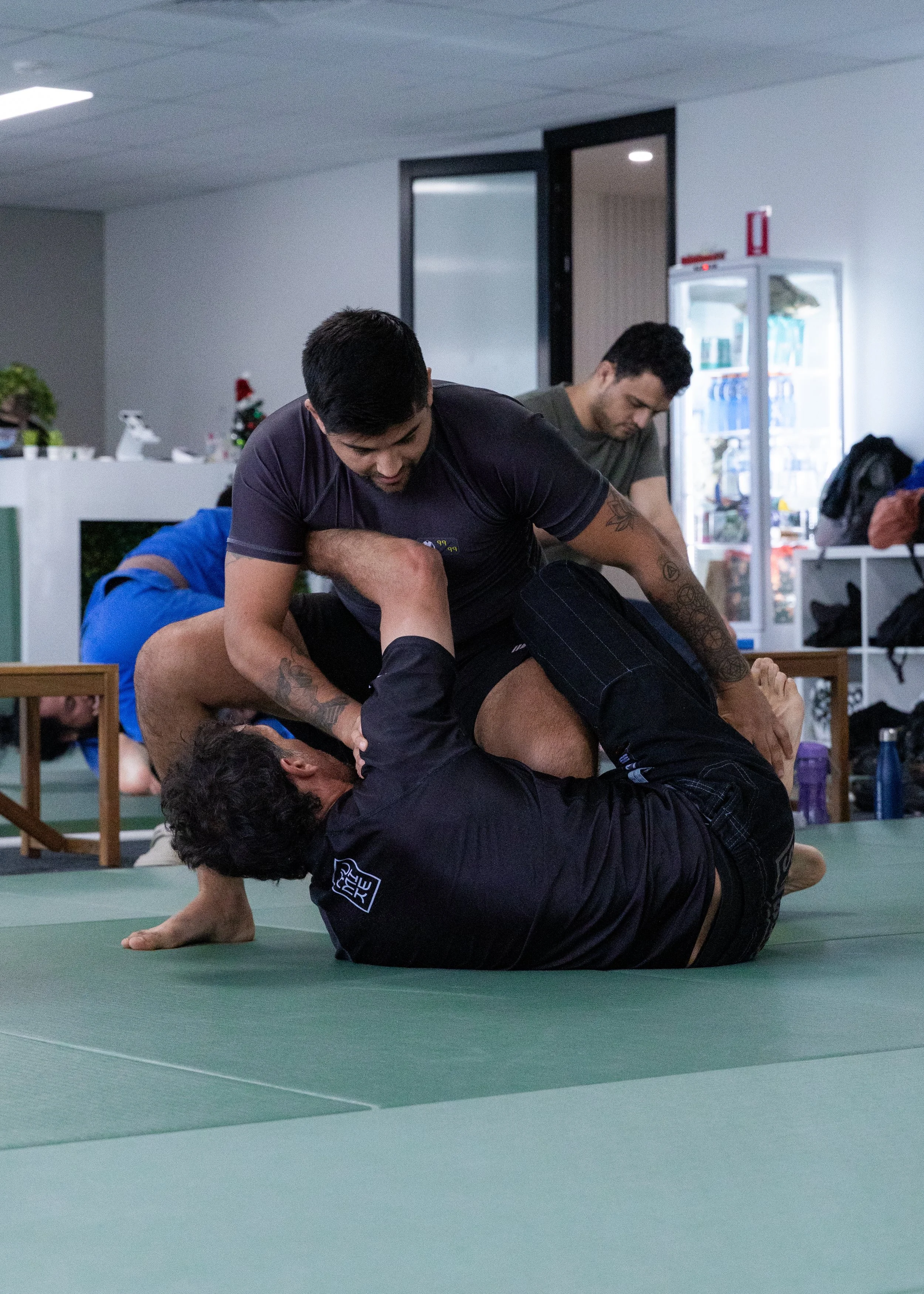Two men practicing Brazilian Jiu-Jitsu on a green mat, with one man in a black gi and the other in casual clothes, engaging in a grappling technique.