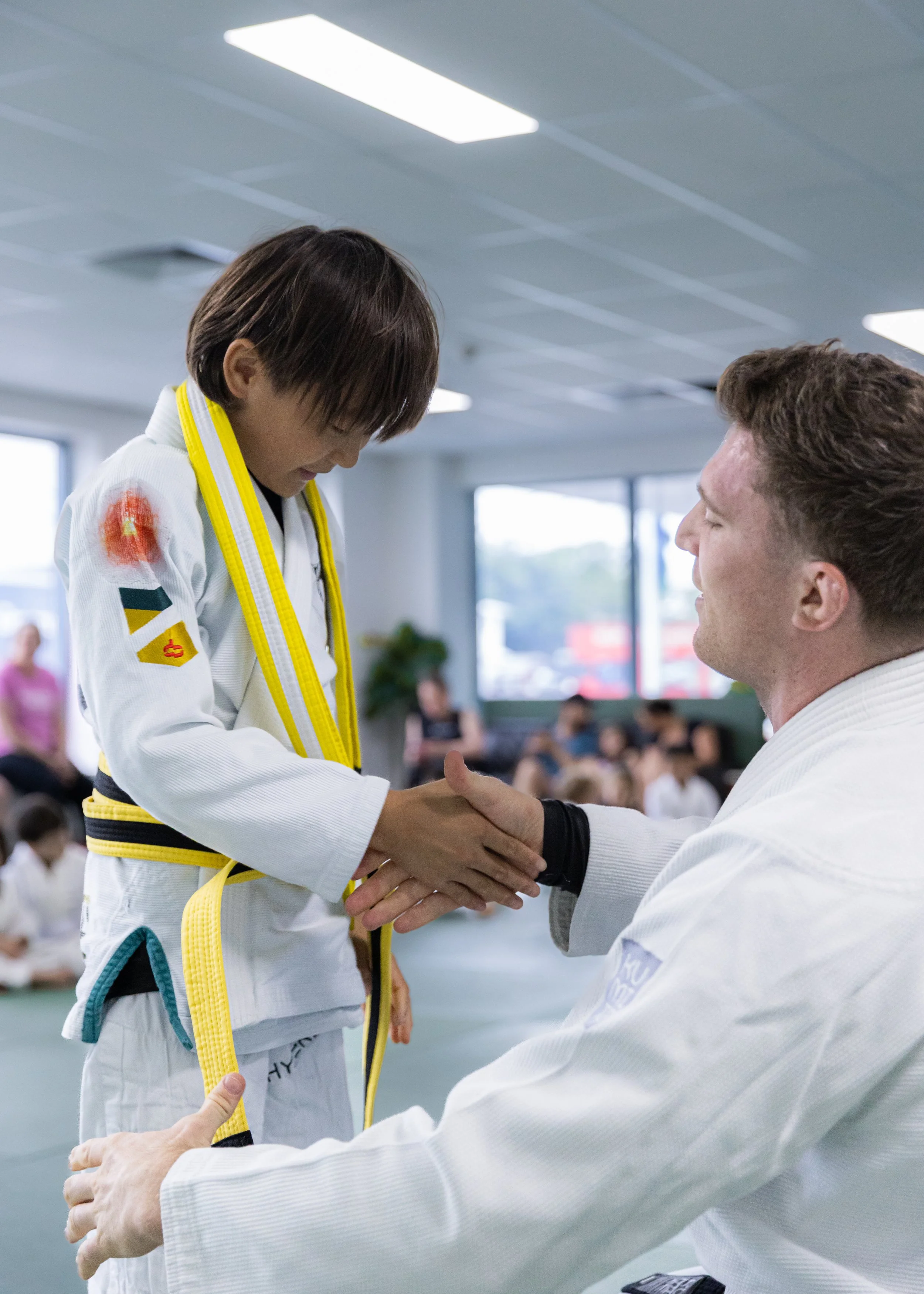 A young martial artist receiving a handshake from an instructor inside a martial arts training facility, with other students and a large window in the background.