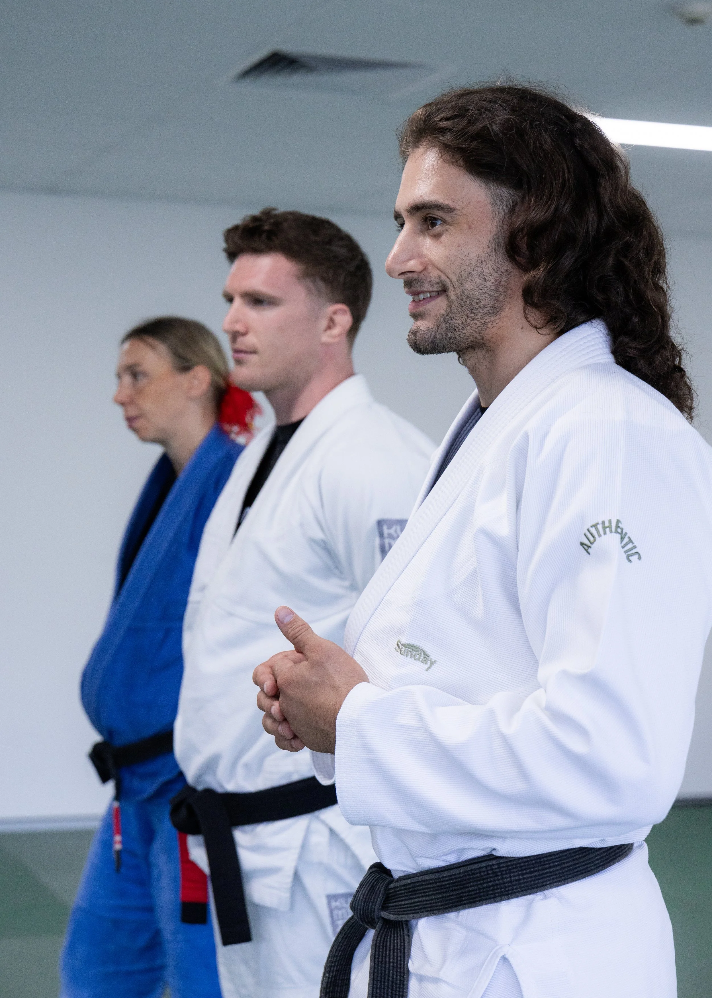 Three martial artists dressed in uniforms with black belts standing in a line, in a dojo or training facility.