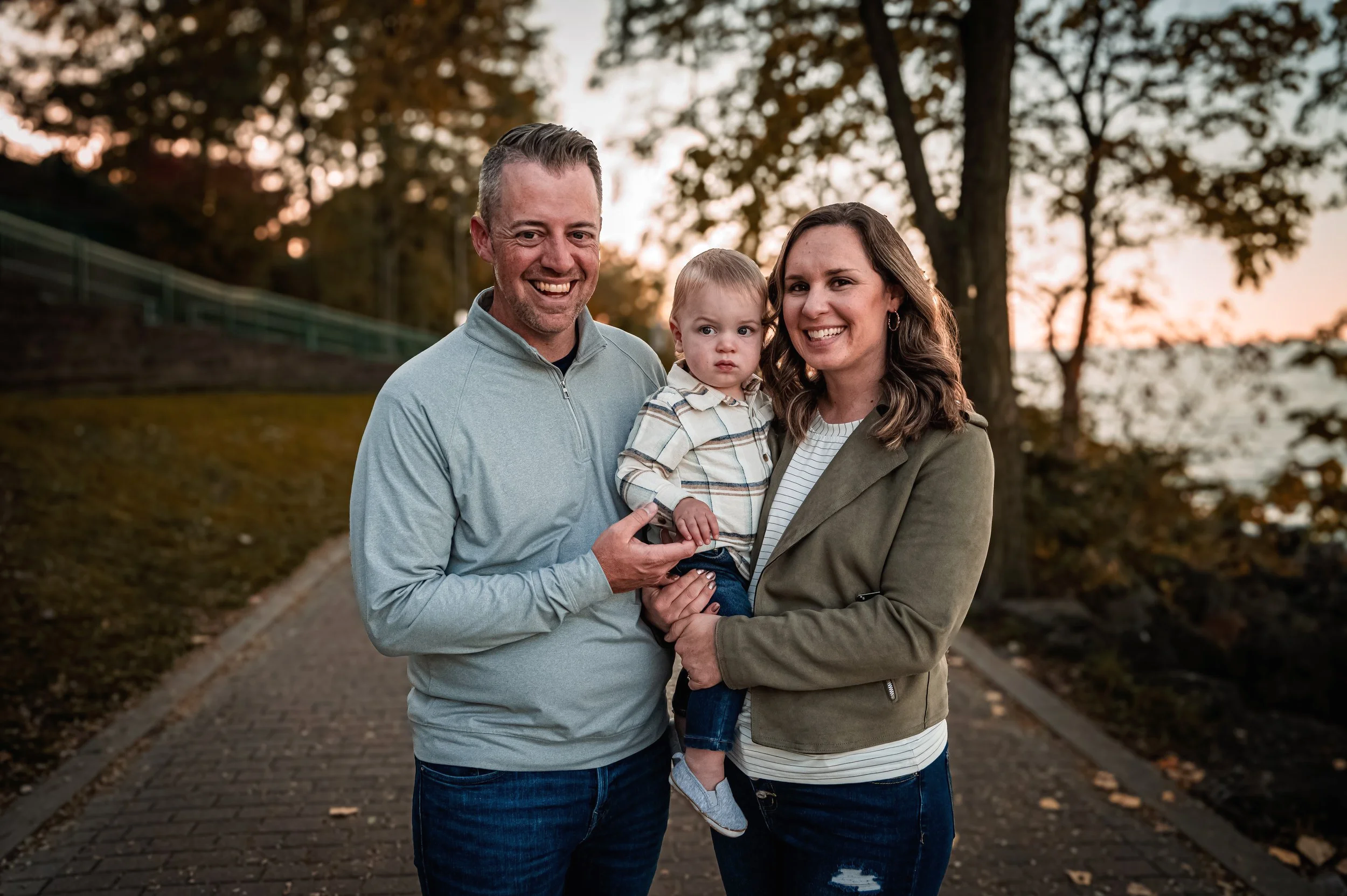 A family of three, a father, mother, and young child, standing on a paved path outdoors during sunset with trees and water in the background, smiling at the camera.