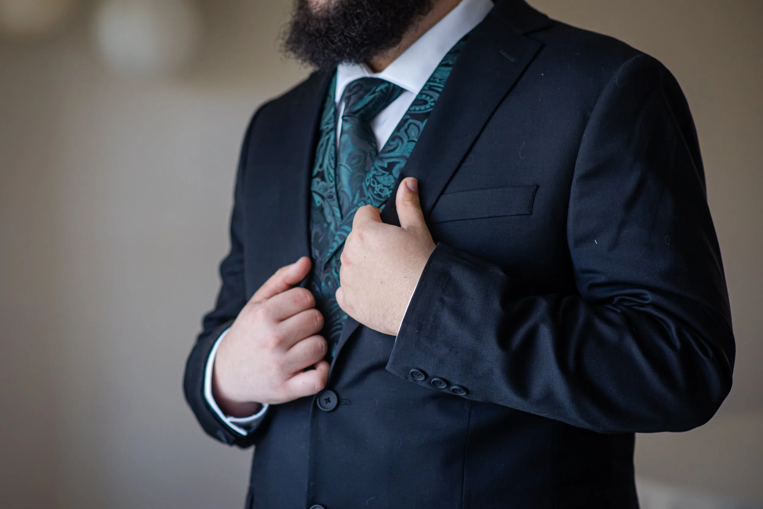 A man in a black suit adjusting his jacket, wearing a teal patterned vest and tie.