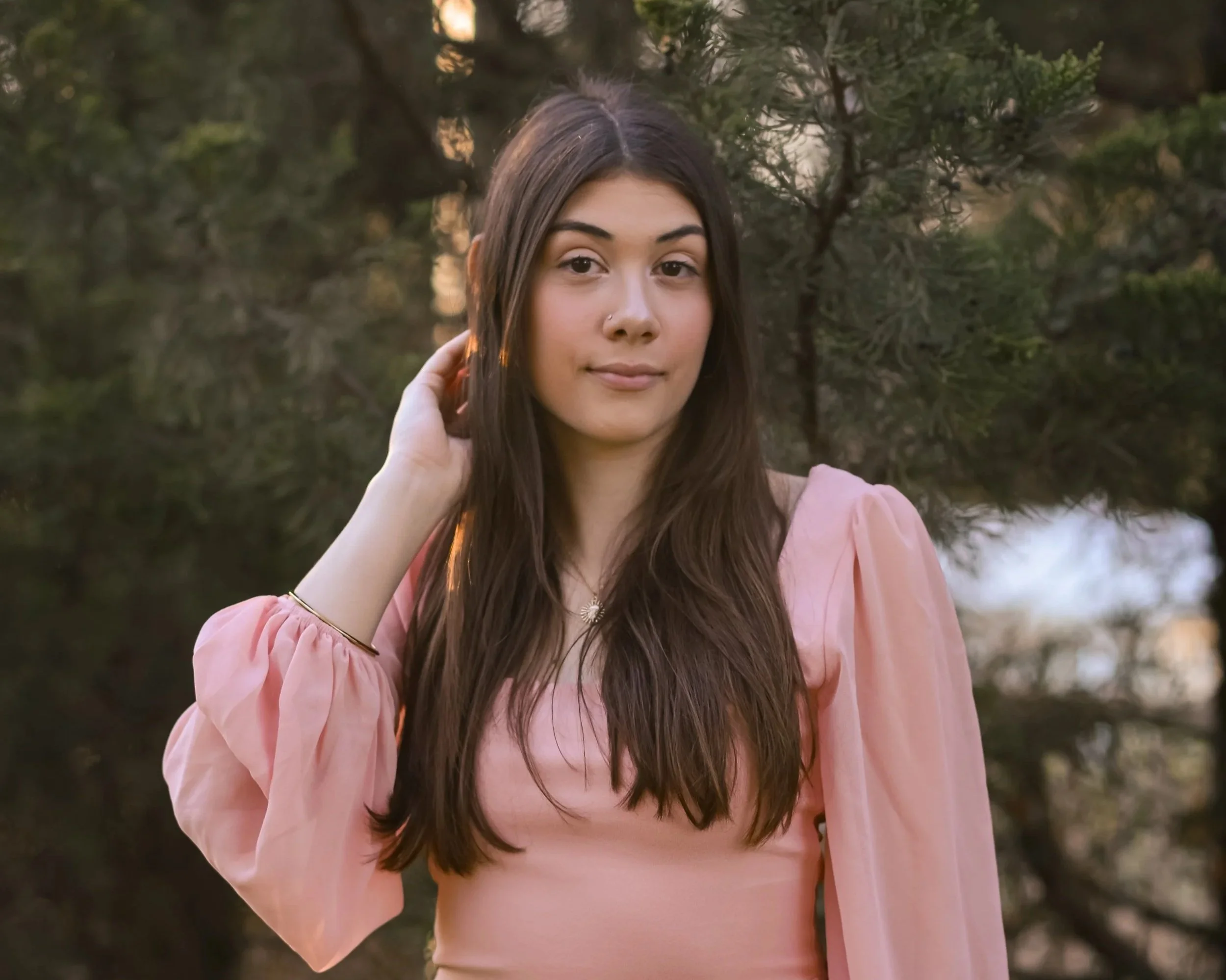A young woman with long brown hair and a nose piercing wearing a pink dress, outdoors with green foliage in the background.