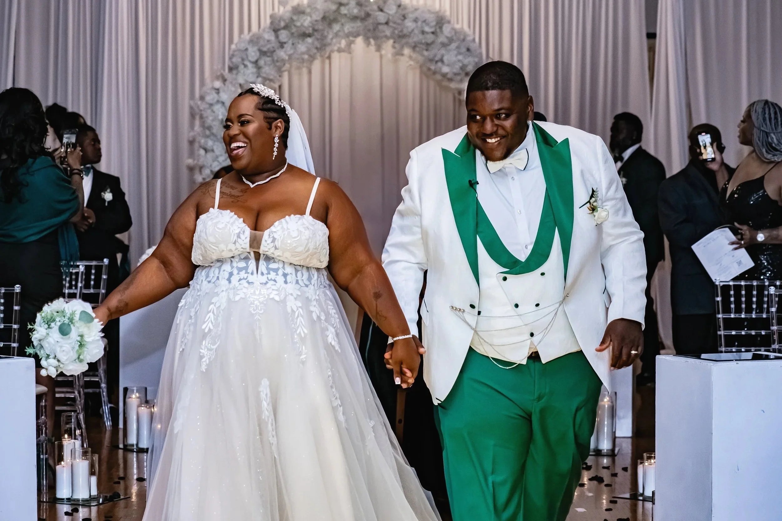 A joyful bride and groom holding hands during their wedding ceremony, dressed in wedding attire, in a decorated venue with guests watching and taking photos.