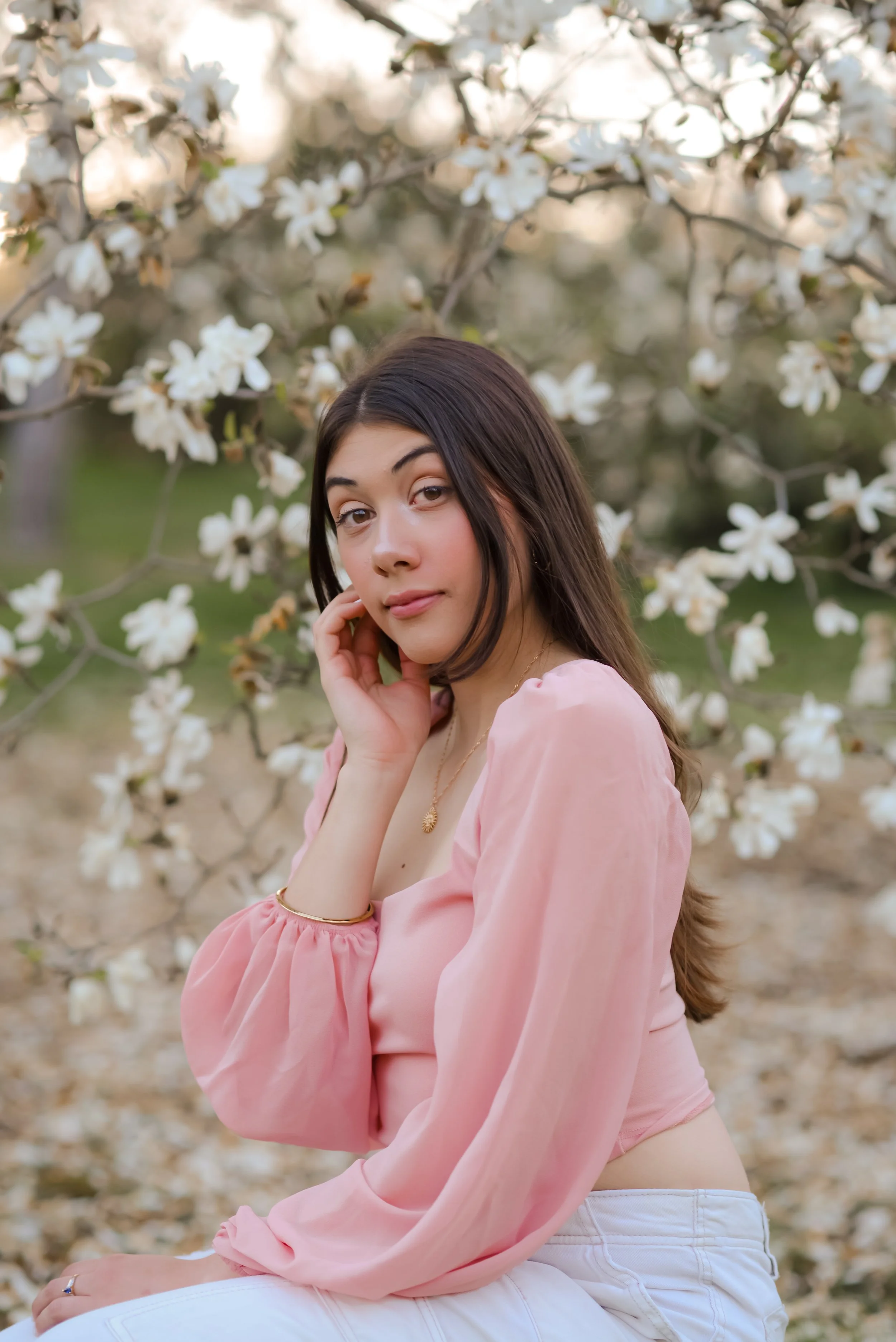 A young woman with long dark hair, wearing a pink blouse and white pants, sitting outdoors in front of blooming white flowers, looking at the camera with a gentle expression.