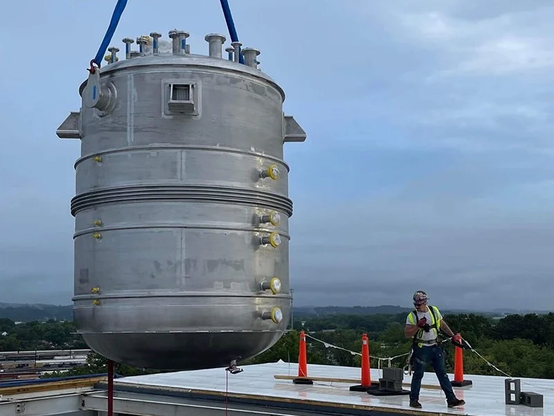 A large industrial metal tank being installed on a building roof, with a worker standing nearby wearing safety gear and holding a tool.