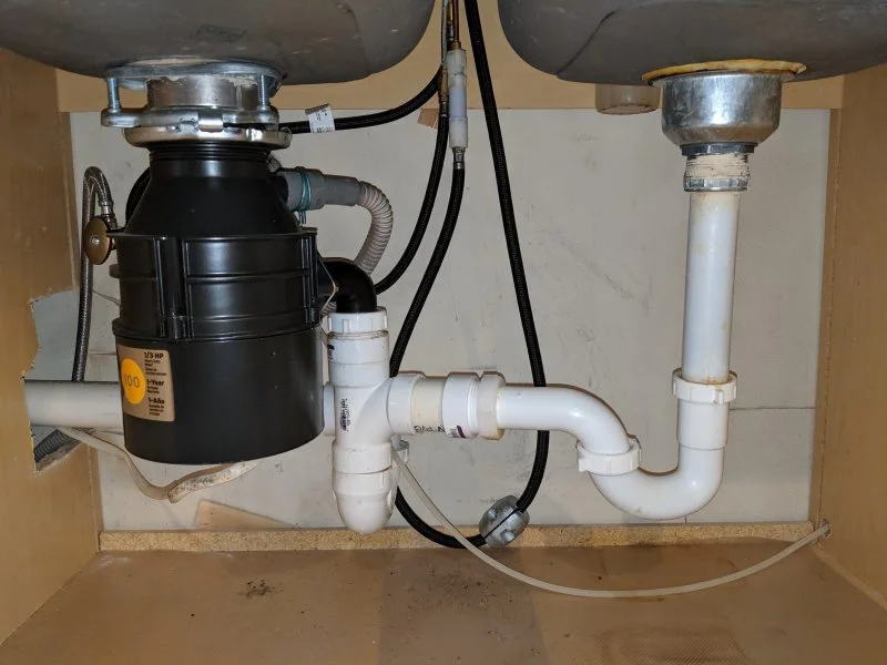 Under sink plumbing with a garbage disposal attached to the drain and connected to PVC pipes, with some black hoses and electrical wiring visible.