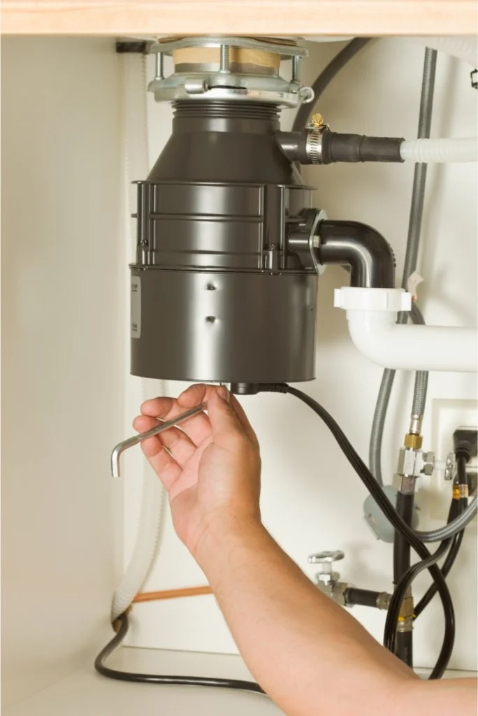 A hand tightening or loosening a pipe fitting under a kitchen sink, with a garbage disposal unit attached to the drain.