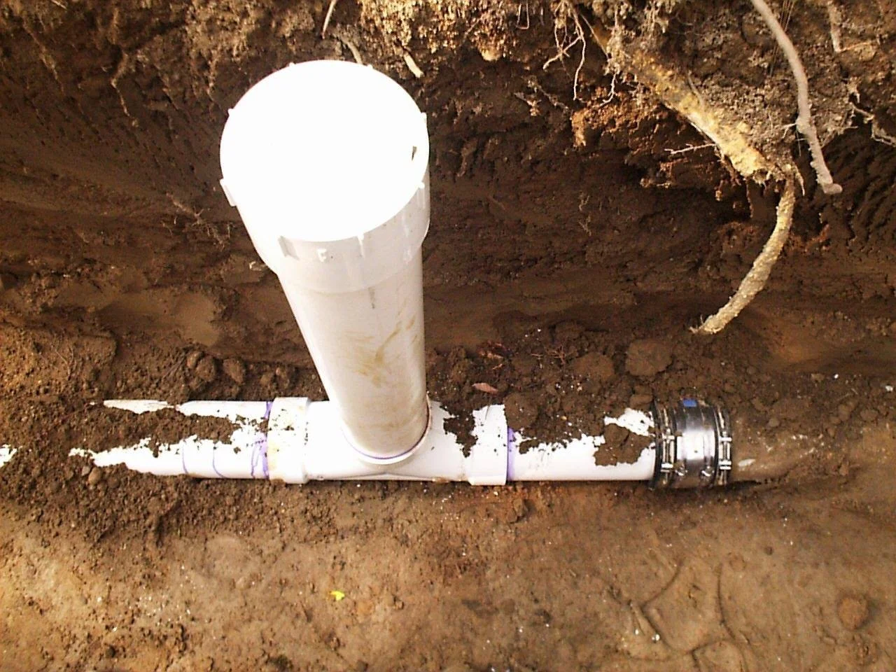 Underground plumbing pipe installation with white PVC pipes and fittings in a dirt trench.