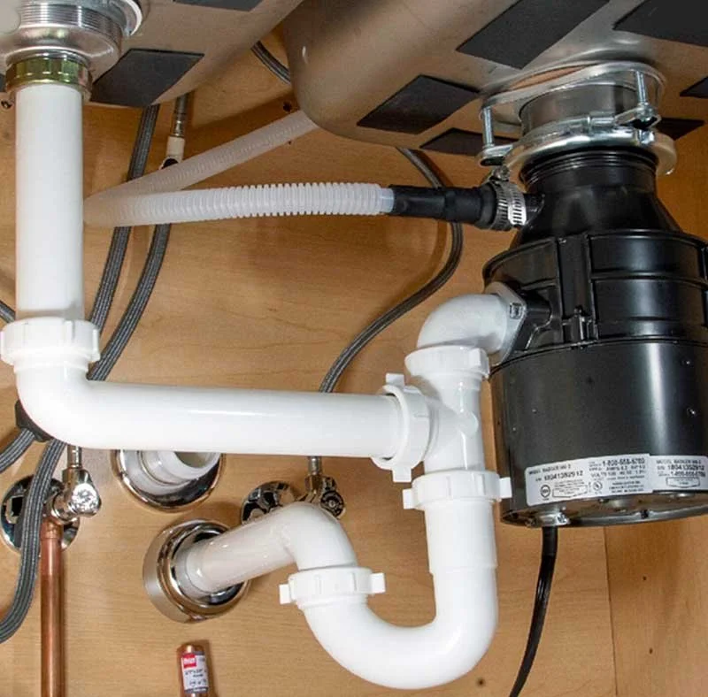 Under-sink plumbing setup with white PVC pipes and a black garbage disposal unit installed under a wooden sink cabinet.