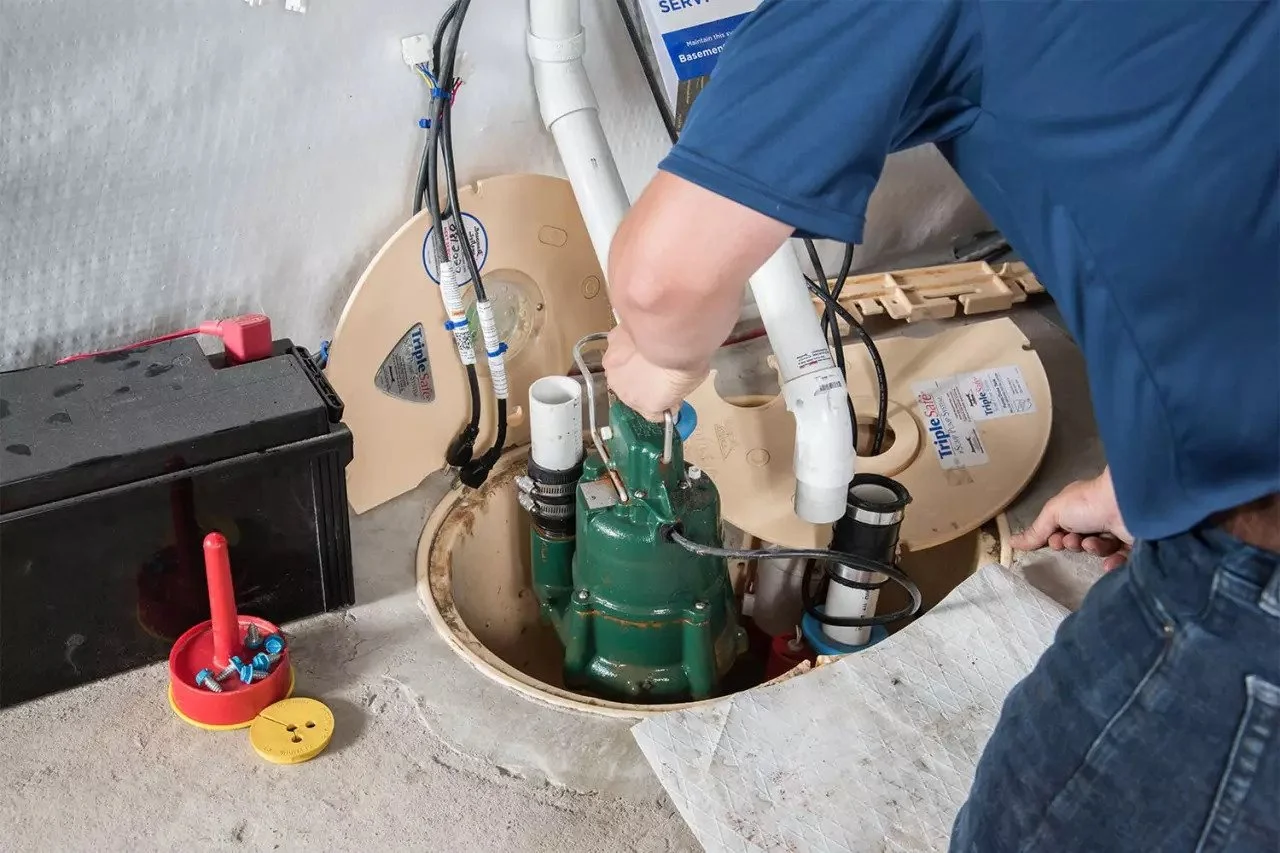 A worker installing or repairing a septic pump in a basement crawl space with plumbing pipes and electrical wiring visible.