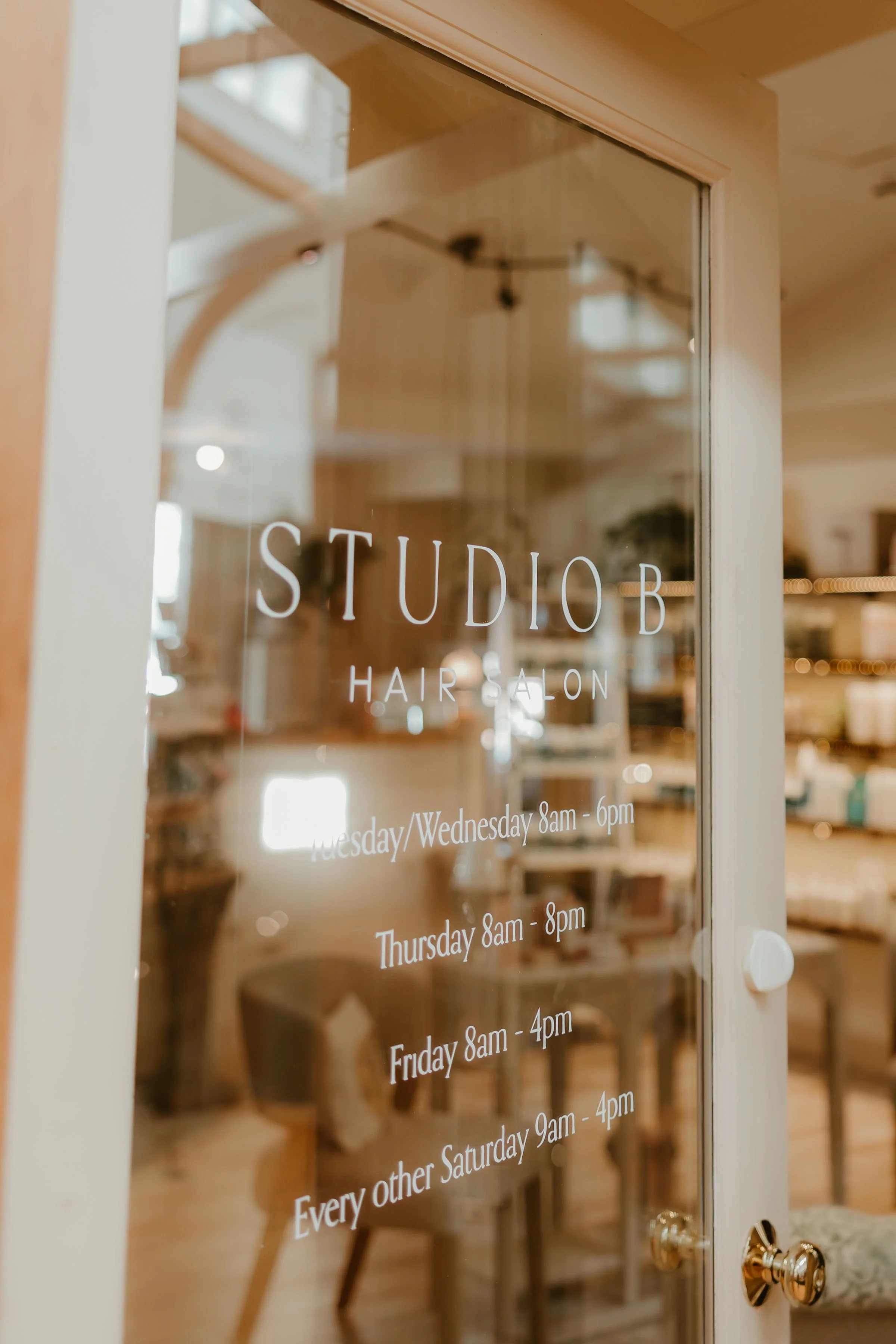 Glass door of a hair salon with white text displaying the salon's name and hours of operation for different days.