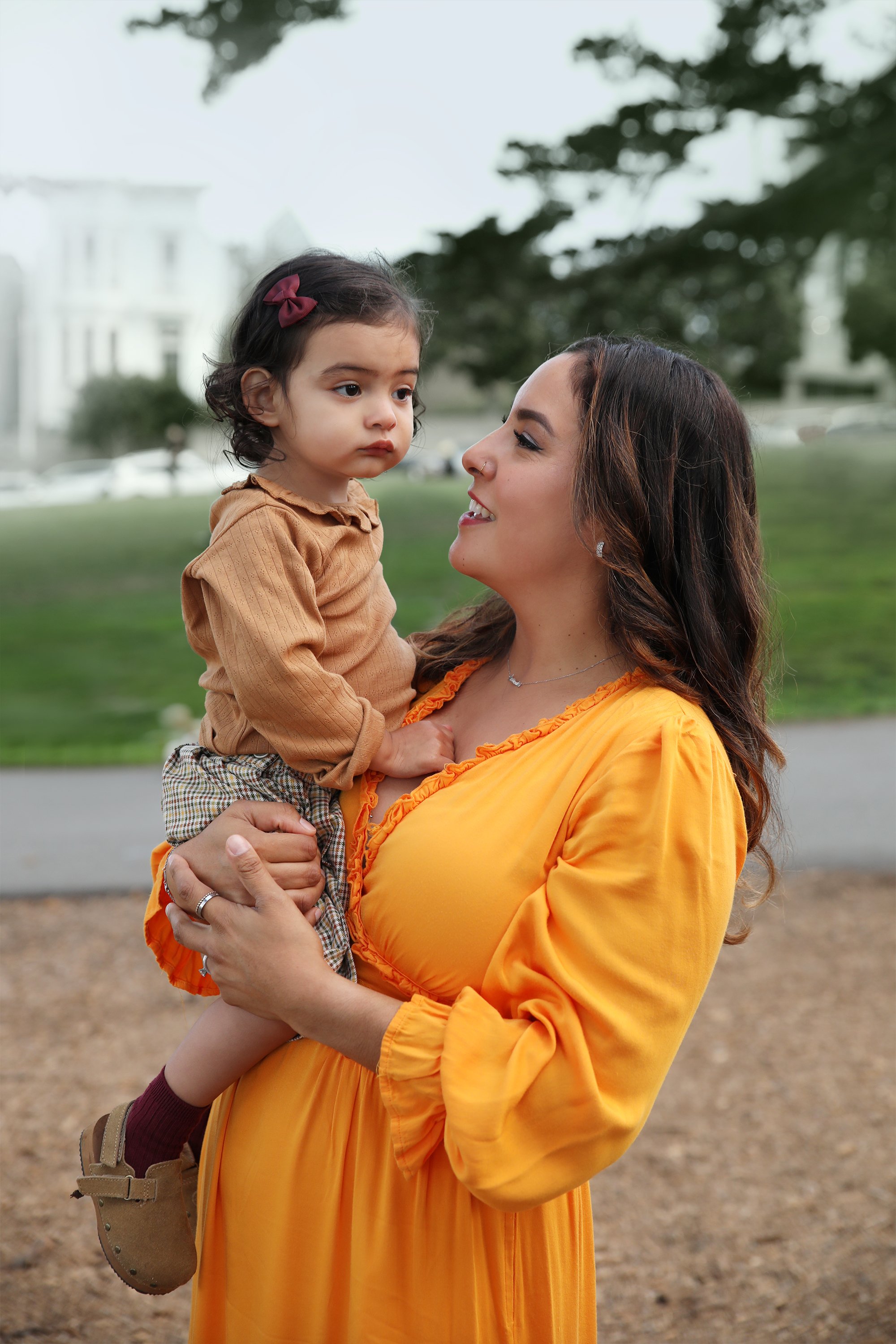 A woman in a bright orange dress holding a young girl in brown and beige clothing, outdoors in a park with trees and blurred buildings in the background.