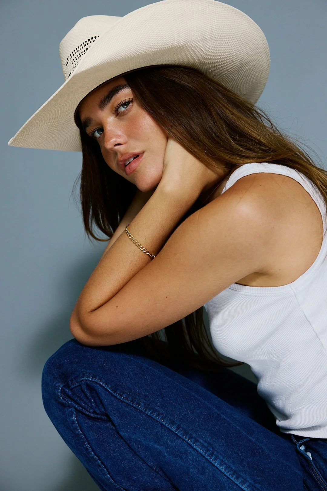 Woman wearing a wide-brimmed white hat, white tank top, and blue jeans, sitting with her knees drawn up and touching her face, against a plain light blue background.