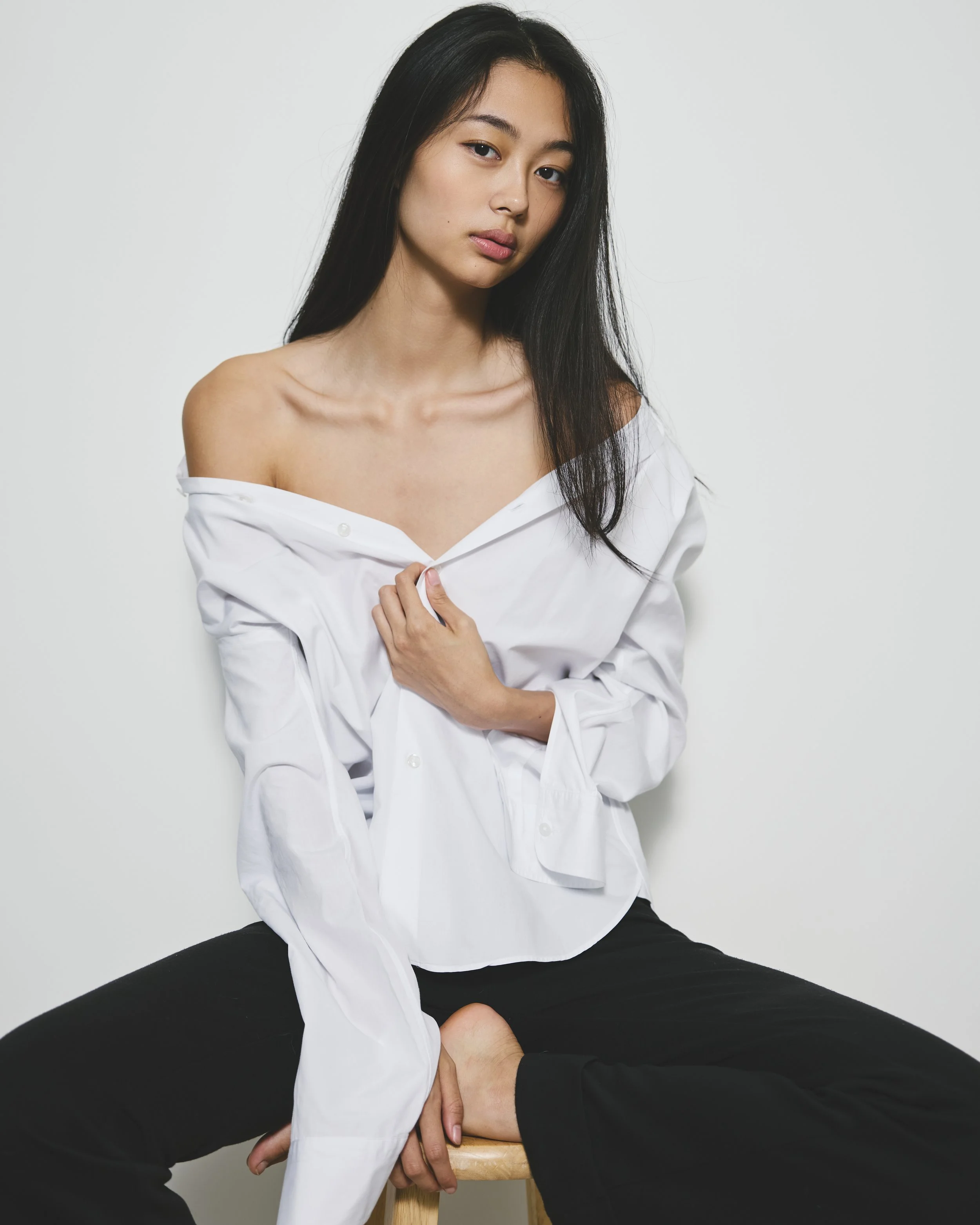 A woman with long black hair, wearing a white off-shoulder shirt, sitting with her legs apart, holding a stool with both hands, against a plain white background.