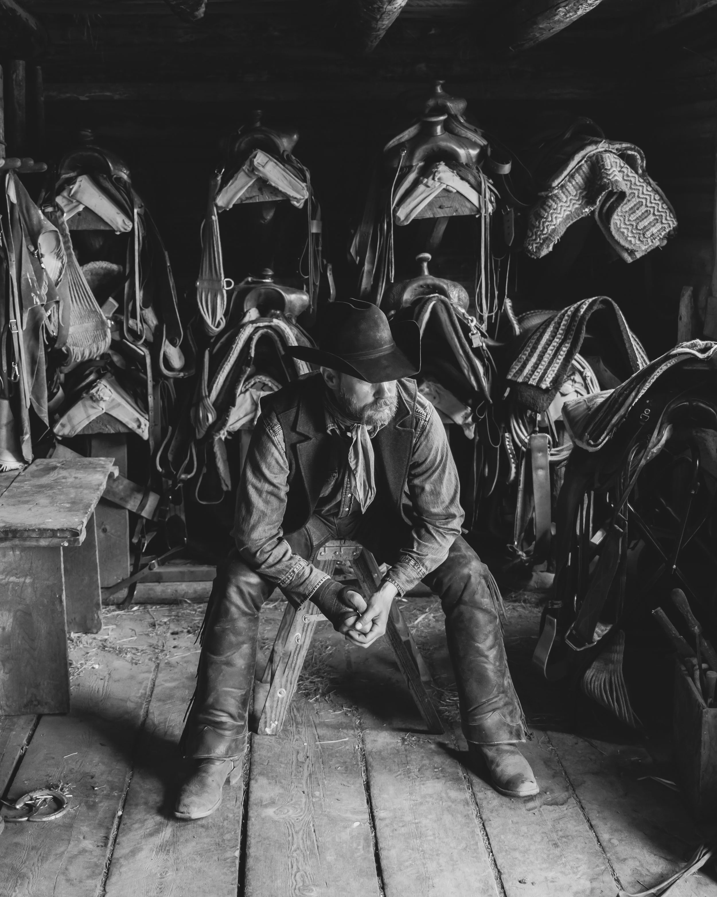 a black and white photograph of a cowboy in a barn sitting on a stool surrounded by saddles