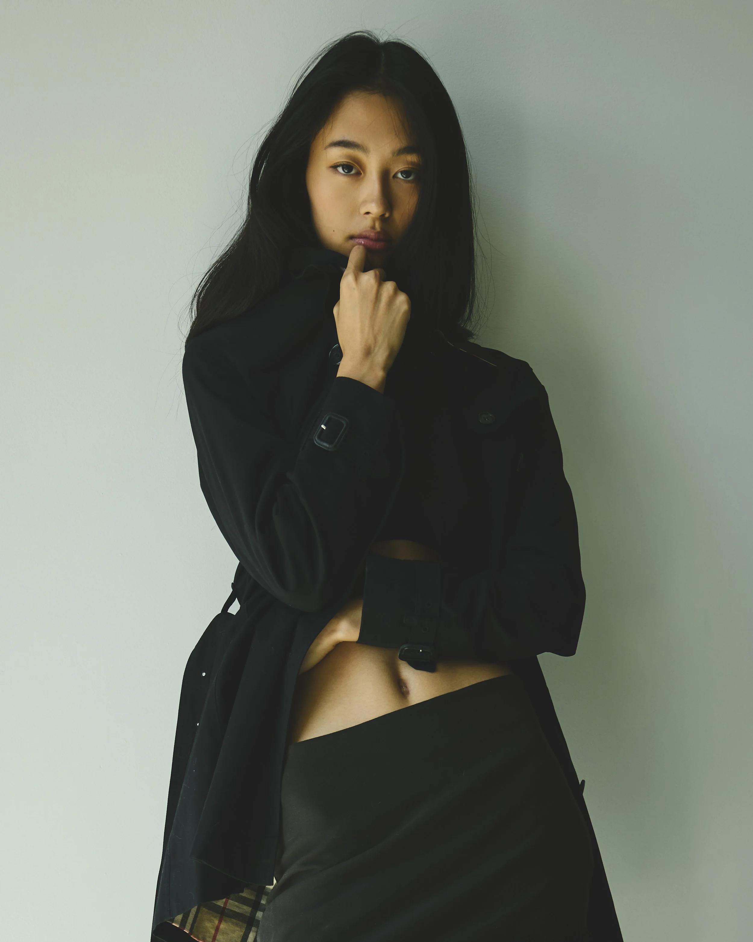 A young woman with long black hair and light skin wearing a black jacket and black skirt, standing against a plain white wall, with her right hand on her chin and her left hand holding the hem of her skirt.