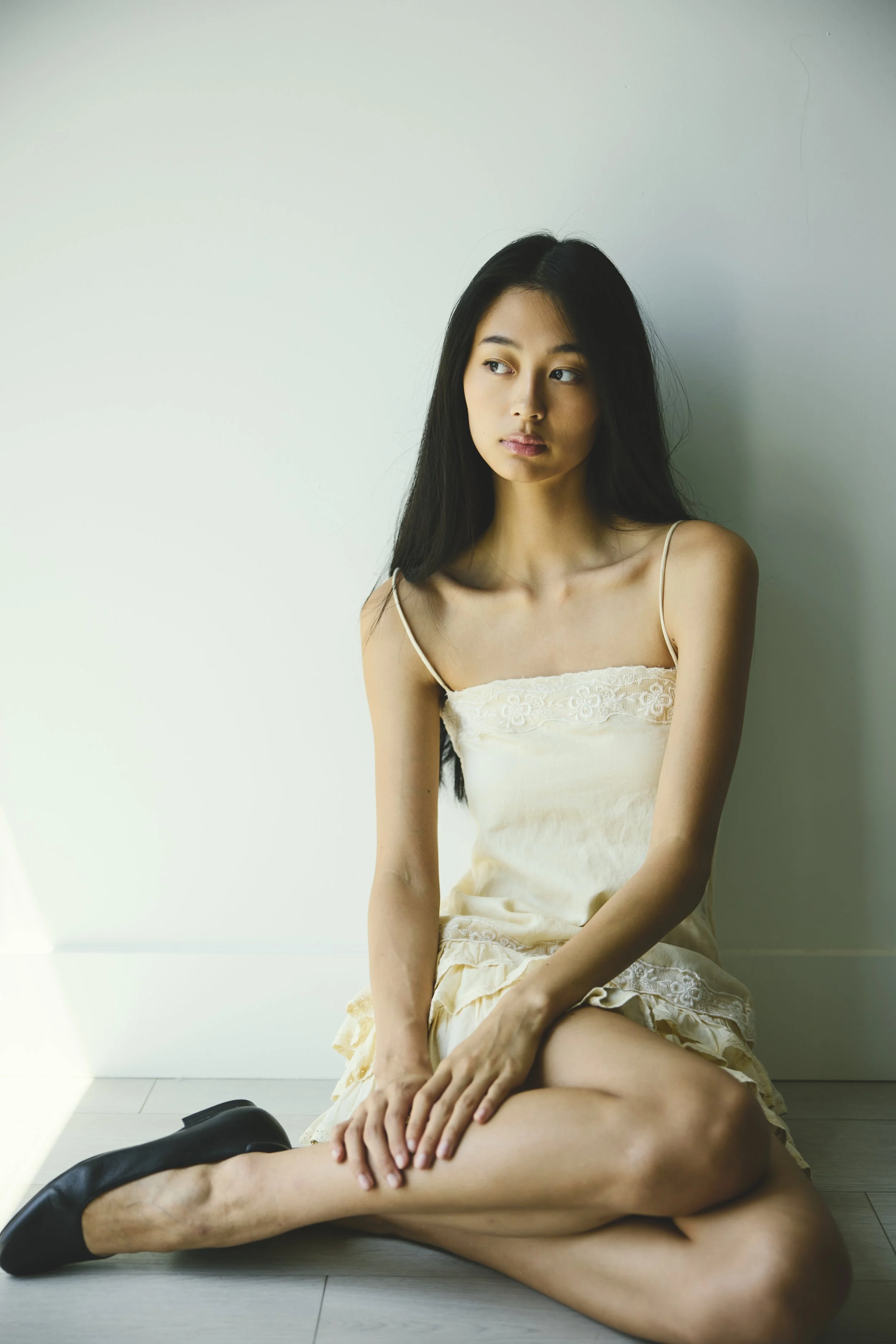 A young woman with long dark hair, sitting on the floor against a plain white wall, wearing a cream-colored dress with lace details. She has a serious expression and is looking off to the side.